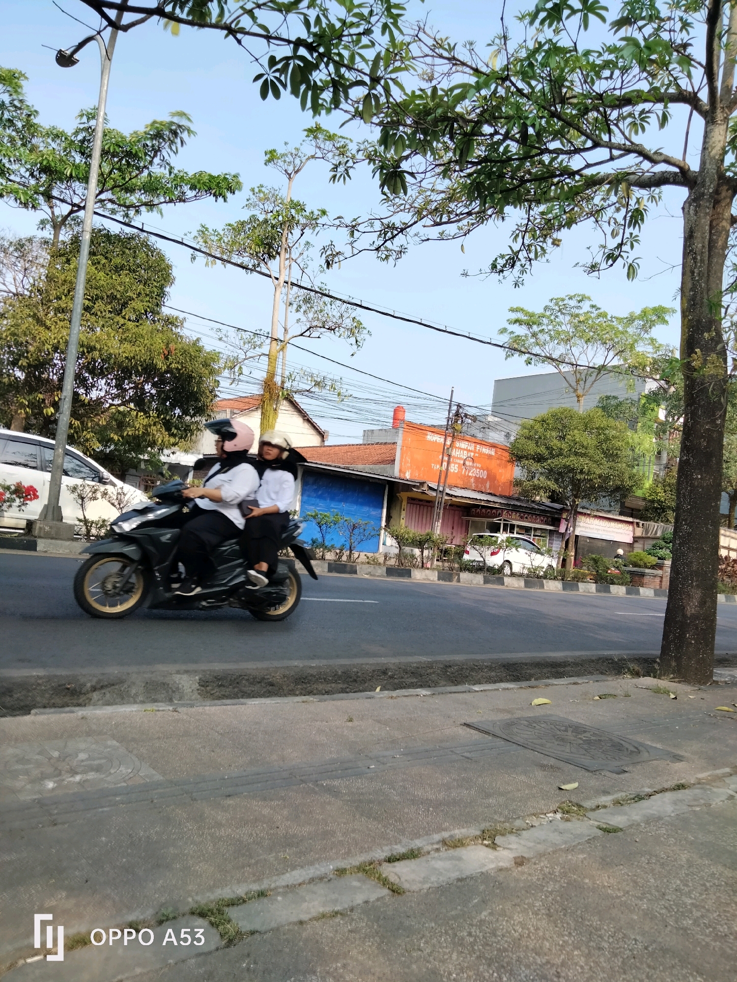 The image depicts a street scene in Kelutan, Indonesia, on a clear, sunny day. In the foreground, a dark-colored scooter carries two individuals, a driver and a passenger, both wearing helmets and modest attire. The scooter is in motion, traversing a paved road. To the left, a white car is visible, parked alongside a sidewalk lined with small trees and bushes. Behind the vehicles, a row of buildings can be seen, some with orange and blue facades, suggesting commercial establishments. The background is filled with lush green trees and a clear blue sky. A lamppost stands on the left side of the road, and power lines are strung across the sky. The overall atmosphere is one of everyday urban life in a tropical setting.