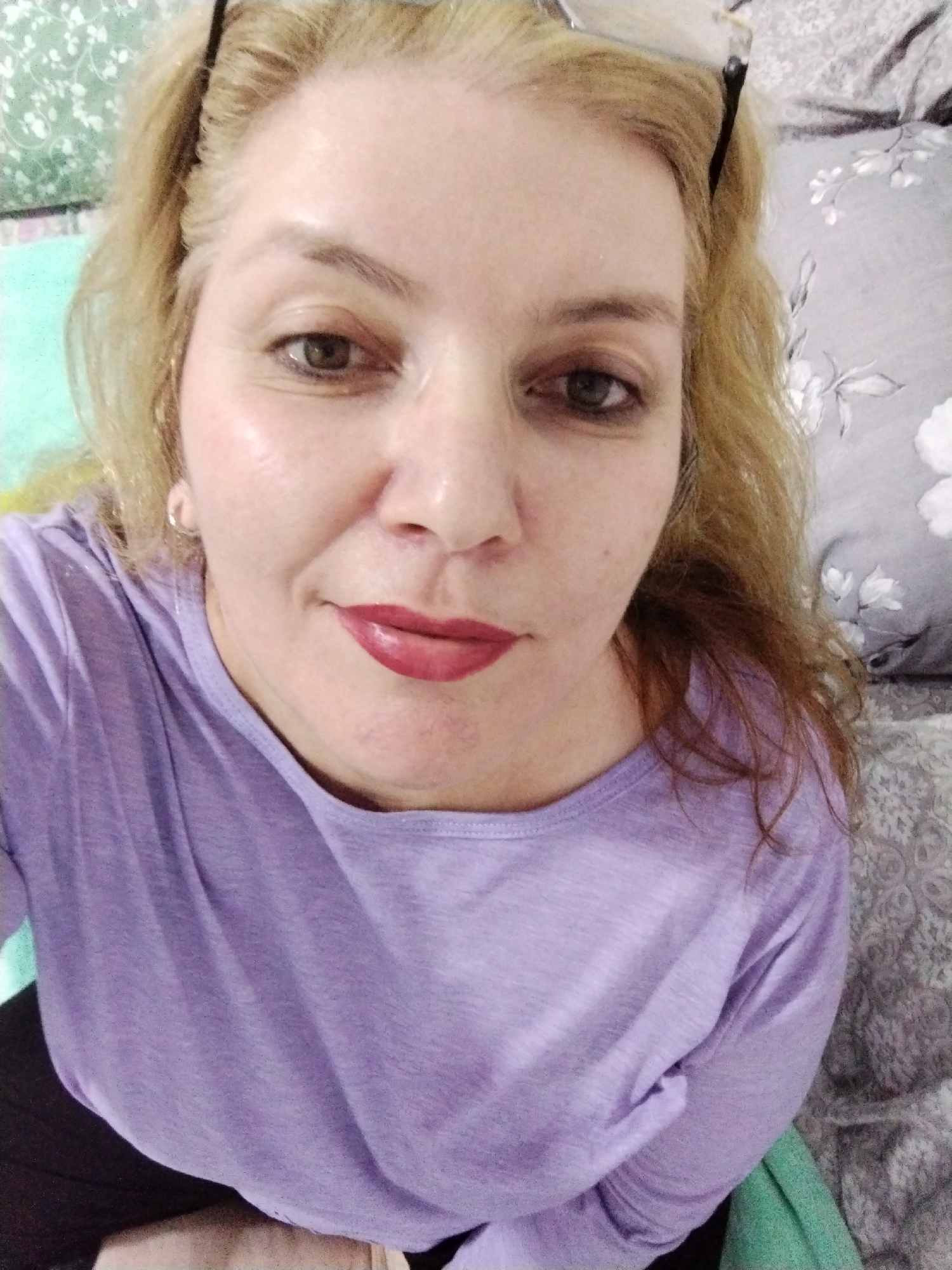 A woman with blonde hair and glasses atop her head is photographed from a close-up, high-angle perspective. She wears a light purple long-sleeved shirt and dark lipstick. Her face is fair with visible skin texture. The background features cushions with varying patterns and colors, including a floral grey cushion. The location and city are impossible to deduce from the visual information.