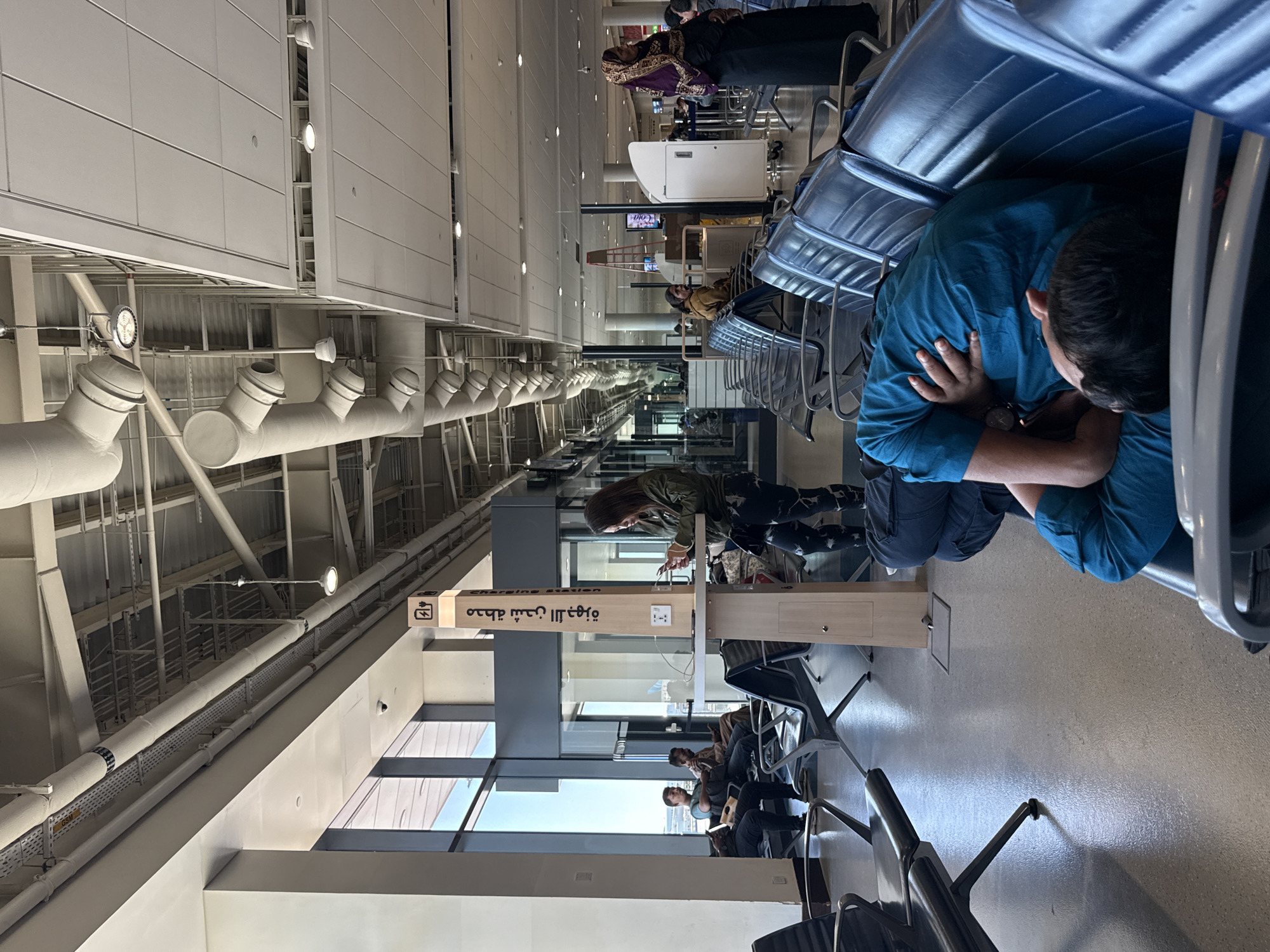 This is a photograph of the interior of an airport terminal in Dubai, United Arab Emirates. The scene depicts a waiting area with rows of blue upholstered seats. In the foreground, a person is sleeping on the seats, with their head resting on their arm. To the right, another person is standing and looking at their phone near a charging station. In the background, other individuals are seated, and some are seen walking. The terminal features large windows that allow natural light to illuminate the space. Overhead, there are exposed pipes, conduits, and lighting fixtures, typical of airport infrastructure. The overall impression is one of a busy but calm public space within the airport.