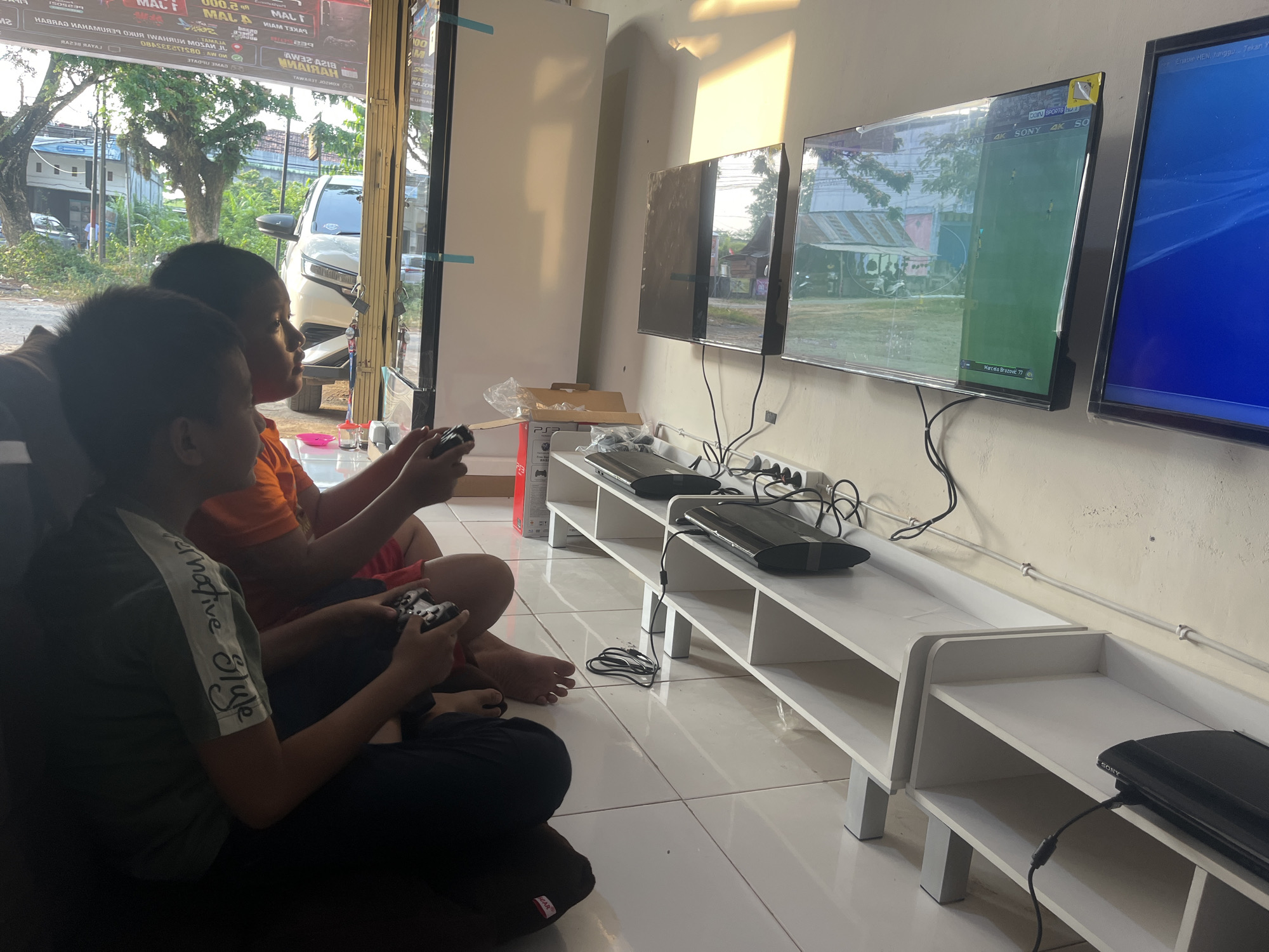 In Sekayu, Indonesia, two boys are playing video games in a brightly lit room, possibly a gaming center, as evidenced by the multiple televisions and game consoles. One boy, wearing a green t-shirt with white lettering on the sleeve, is seated and holding a black video game controller. He is looking to his right, towards the televisions. Beside him, another boy in an orange shirt is also seated and holding a controller, his gaze directed forward. They are positioned in front of a white, multi-tiered shelving unit where two black Sony PlayStation 3 consoles are placed. Three large televisions are mounted on the wall above the shelves, displaying video game content. The room has a tiled floor and large windows that offer a view of trees and a white car parked outside, suggesting an urban or suburban setting. Sunlight streams in from the right, casting a warm glow on the wall. Various cables and power outlets are visible along the wall and on the shelves, indicating the setup for the gaming equipment. A crumpled plastic bag and a cardboard box are also present on the floor near the shelves.