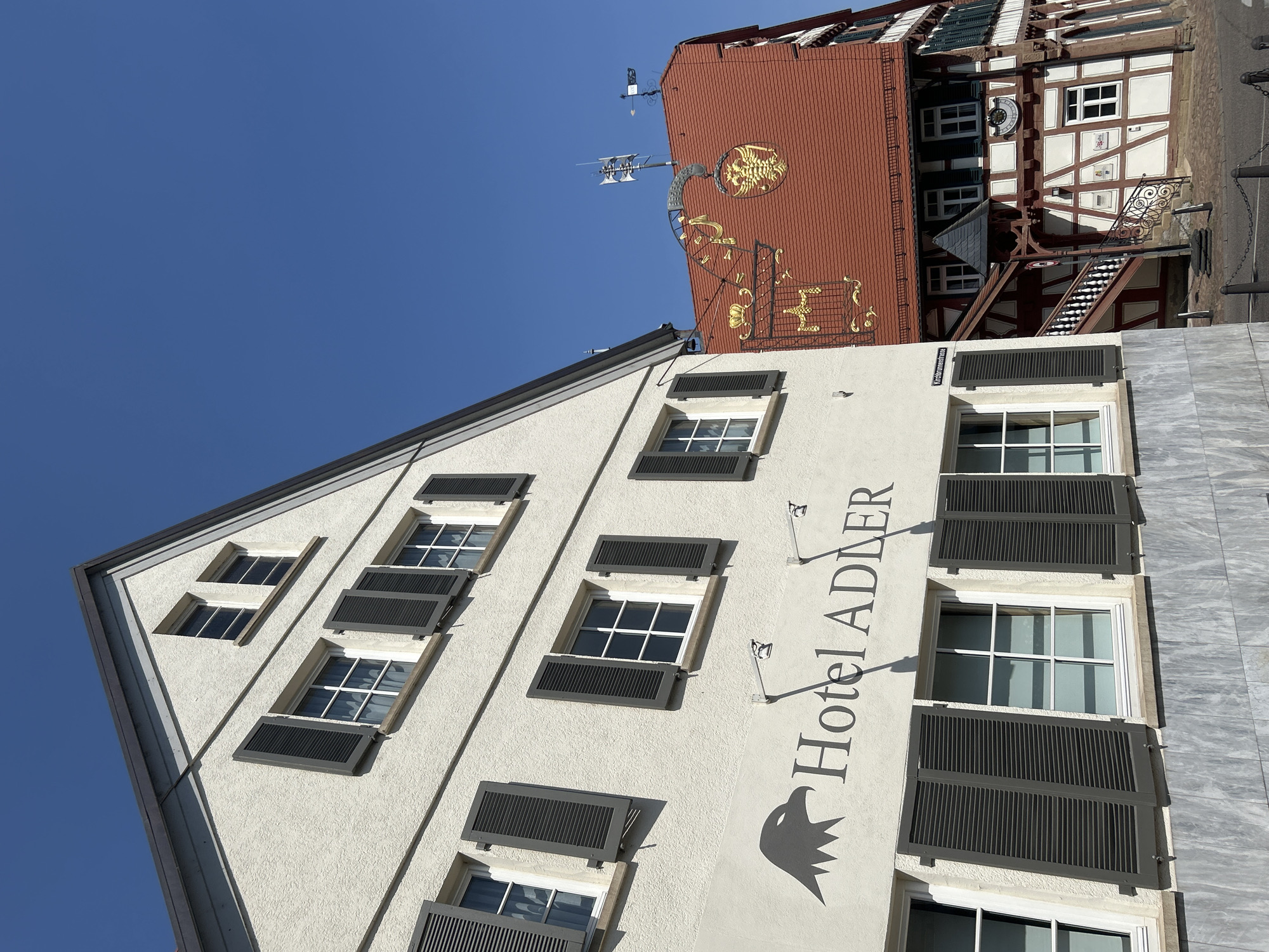 The image depicts a street view in Bad Friedrichshall, Germany, featuring two traditional buildings. The building on the left is a white, multi-story structure with a gabled roof and multiple windows, some of which are adorned with dark shutters. A prominent sign on this building reads "Hotel ADLER" and features a stylized image of an eagle. The building on the right is a half-timbered structure with a red tiled roof. It has a decorative golden crest with an eagle motif above its entrance, which is accessed via a red staircase with white railings. The sky above is clear and blue.
