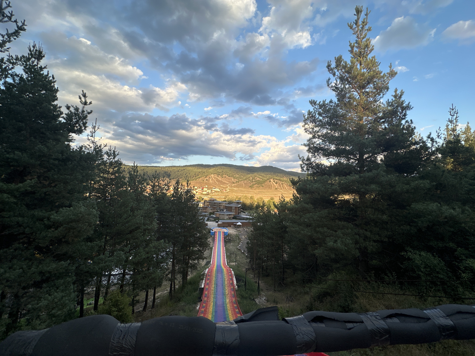 The image shows a rainbow-colored slide extending down a grassy slope, surrounded by pine trees. In the background, a small village with wooden buildings is nestled in a valley, with rolling hills beyond. The sky is partly cloudy, with the sun casting a warm glow on the landscape. The perspective suggests the photo was taken from a higher vantage point, possibly at the top of the slide or an adjacent structure. The overall scene depicts a recreational area in a mountainous region, with the slide appearing to be a central attraction.