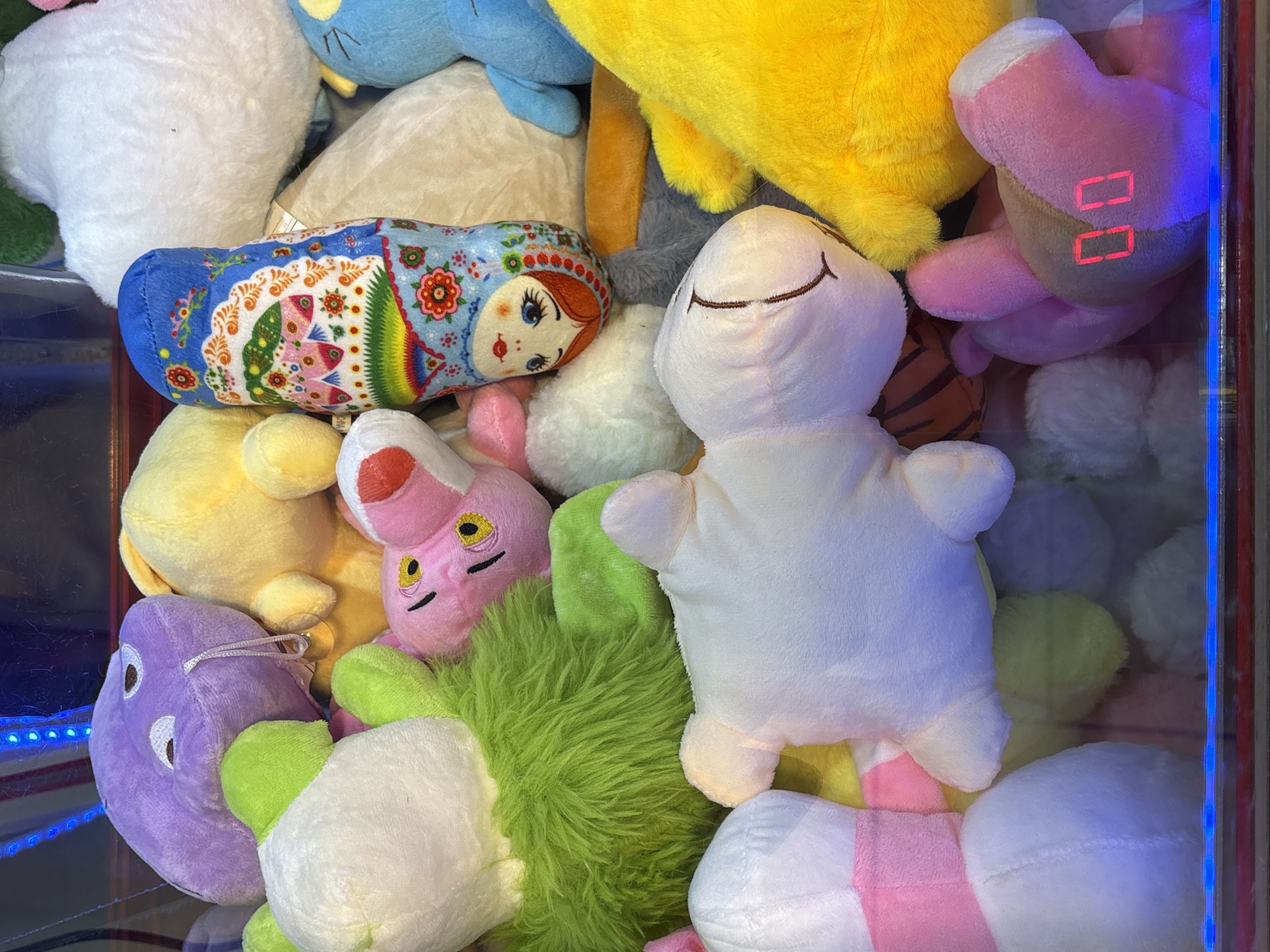 The image depicts a collection of plush toys inside a claw machine. Toys include a blue-and-red matryoshka doll, a white animal with stitched eyes, a pink bunny with yellow eyes, a yellow plush toy, and a pink plush heart with red digital numbers on it. A purple plush toy with white eyes and a green toy with fluff also appear. The toys appear to be jumbled together. The scene seems to be inside a claw machine, given the blue lights and clear case. There's no evidence in the image that definitively locates it in Birobidzhan, Russia.