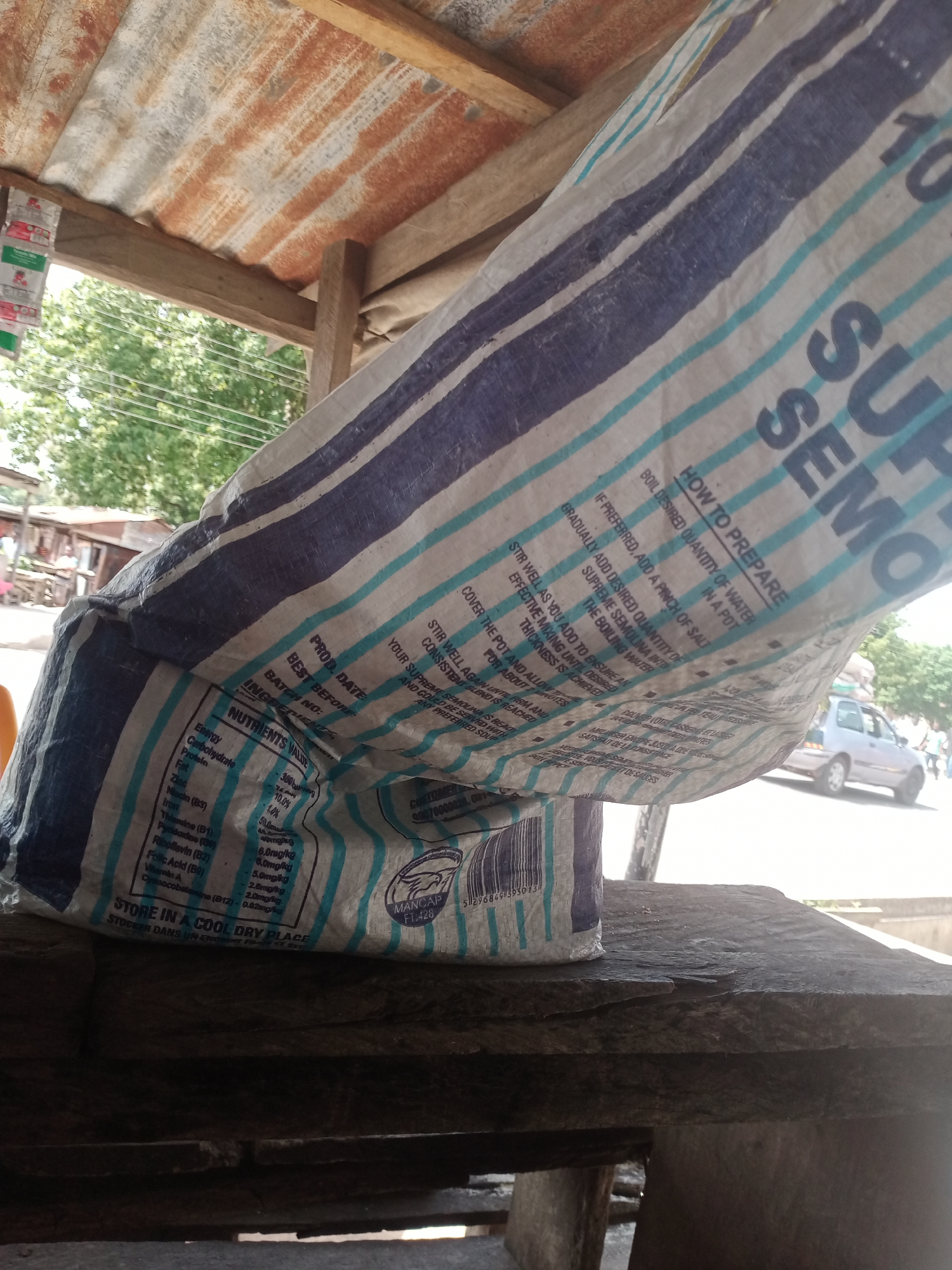 This media file shows a close-up of a large, striped bag of semolina, likely in a market setting. The bag, predominantly white with blue and teal stripes, is leaning against a rustic, weathered wooden surface. The text on the bag indicates it is "SUPREME SEMOLINA" and provides nutritional information, preparation instructions, and storage advice in English and French. A barcode is also visible.
In the background, a glimpse of a street scene in Jalingo, Nigeria, can be seen. A light-colored car is parked or driving past, and the presence of trees and structures suggests an outdoor environment, possibly under a tin-roofed shelter. The lighting indicates it's daytime, with shadows suggesting bright sunlight. The overall impression is of a local market stall or a small shop. No people are clearly visible or identifiable in the image, with the focus being on the product and its packaging.