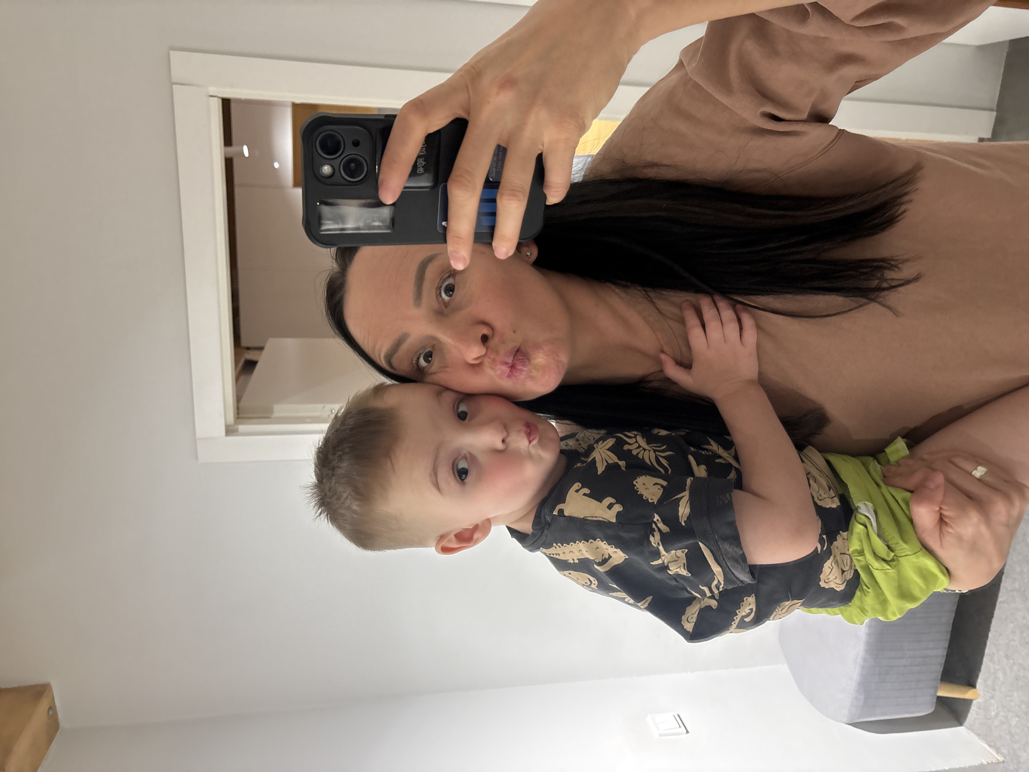 An indoor mirror selfie depicts an adult female holding a young male child. The female, possessing long dark hair, wears a light brown long-sleeved garment. She holds a black smartphone, equipped with a multi-lens camera and a black protective case featuring a blue-striped cardholder, in her left hand to capture the reflection. A silver-colored ring is present on her left ring finger. Her eyes are wide, and her lips are pursed forward. The child, estimated to be between three and five years old with short light brown hair, wears a dark t-shirt adorned with light-colored patterns and bright green shorts. He is held by the female, with his head leaning against her left cheek, and also exhibits a pursed-lip expression. A small hand, identified as the child's, rests on the female's chest. The background consists of plain light-colored walls. A grey upholstered bench with light wooden legs is positioned in the lower-left portion of the frame. A white-framed doorway is centrally located in the background, revealing a room with recessed ceiling lights beyond. A light-colored wooden beam is visible in the upper-left ceiling area, and a white rectangular fixture, possibly a light switch, is on the left wall. The specific location within the city of Chaikovsky, Russia, is not ascertainable from the visual information provided.