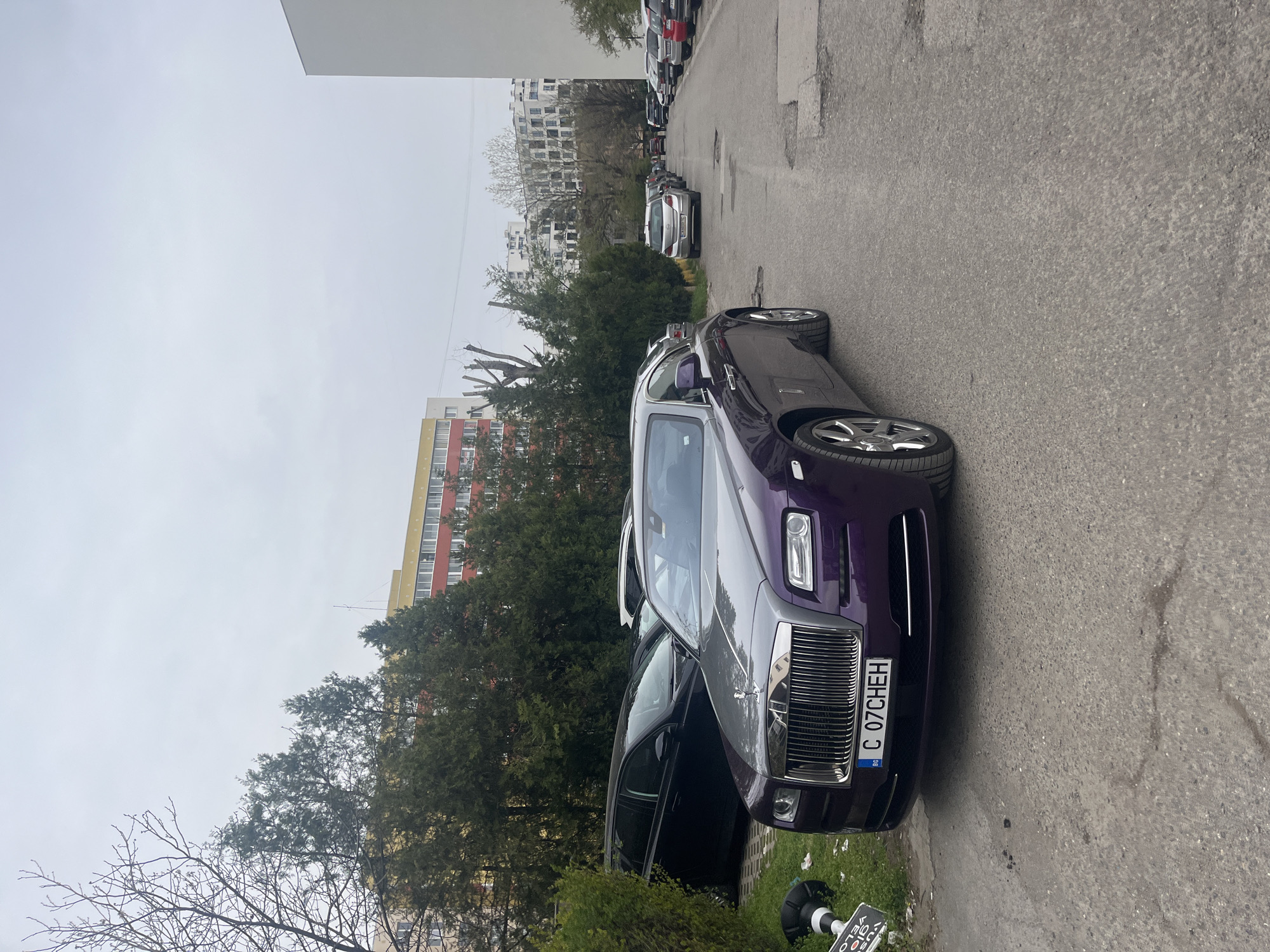The image shows a two-tone (silver and purple) Rolls-Royce Ghost parked on a narrow street in what appears to be a residential area. The license plate reads "C 07CHEH," indicating that the car is registered in Sofia, Bulgaria, given that "C" is the vehicle registration code for Sofia. The area features residential buildings, some with yellow and red facades, and mature trees. The sky is overcast. Several other vehicles are parked further down the street. A traffic sign is visible on the right edge of the road. The door of the Rolls-Royce is open on the passenger side. No people are visible in the immediate vicinity of the car. The photo suggests a quiet, urban setting.