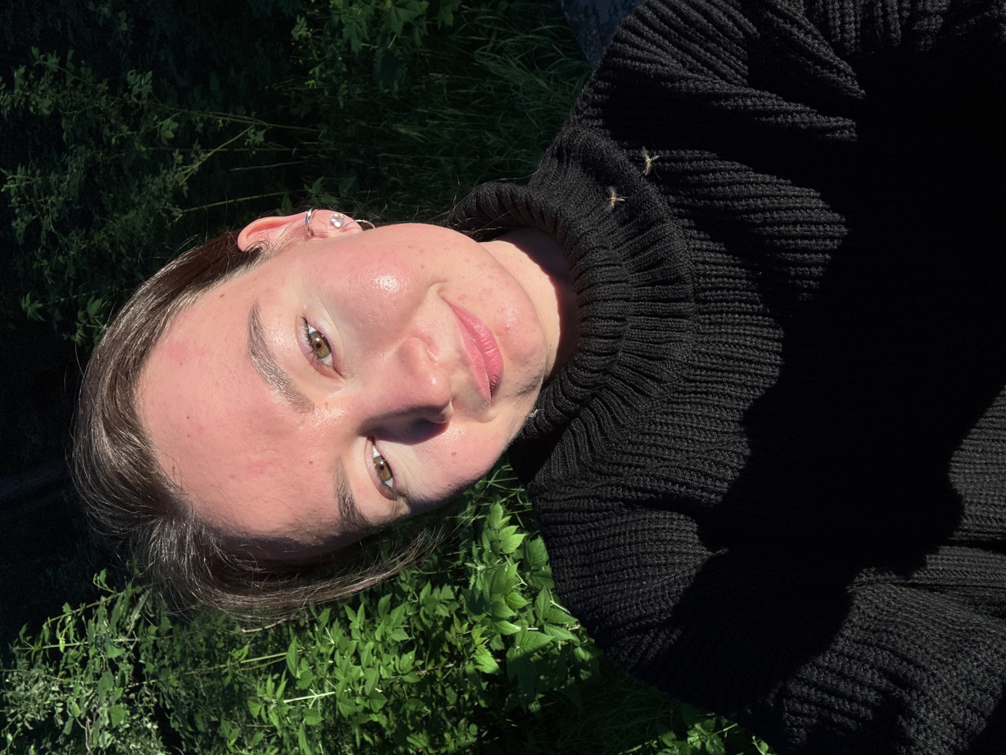 The image is a close-up portrait of a young person, possibly female, positioned outdoors amidst lush green foliage. The subject is looking directly towards the camera with a soft, relaxed expression, characterized by a slight smile and a calm gaze. Their dark hair frames a fair-skinned face, which shows some natural redness or blush on the cheeks. Brown eyes are visible, and the left ear features small silver-toned earrings, including a hoop and a stud with a clear, sparkling stone.
The person is wearing a thick, black knitted sweater with a high collar, the ribbed texture of which is clearly visible and accentuated by the lighting. Small, light-colored details, possibly decorative knots or loose threads, are sparsely visible on the sweater, catching the light.
The background and foreground are dominated by vibrant green leaves and dense plant life, suggesting an overgrown natural setting such as a garden, field, or forest edge. Bright sunlight illuminates parts of the subject's face and the surrounding foliage, creating strong highlights and distinct shadows. This indicates a sunny daytime scene, likely late morning or afternoon. The overall atmosphere conveyed is serene and natural. The image is set in Chaikovsky, Russia. There is no visible text in the media file.