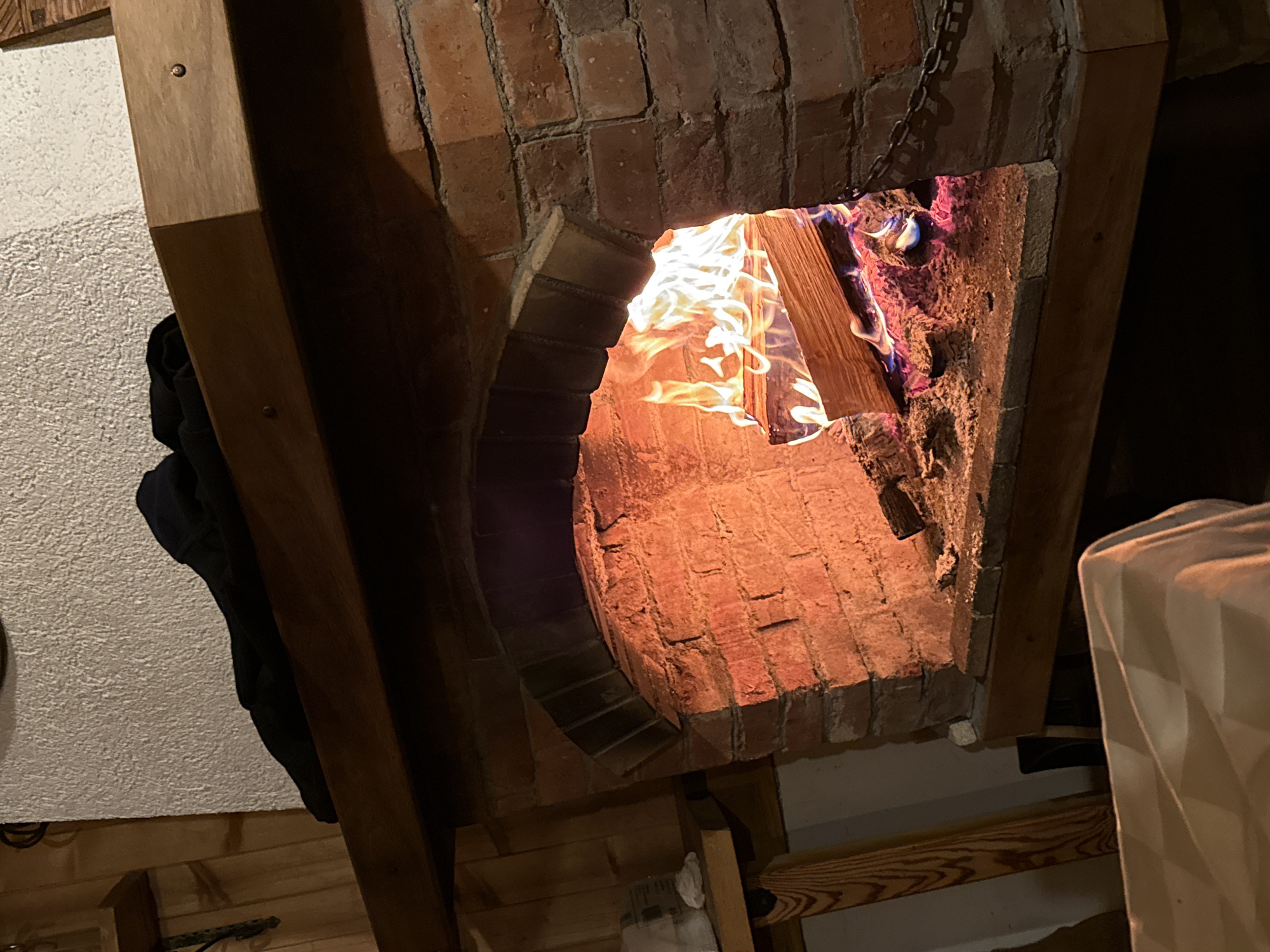 The image shows a brick fireplace with a fire burning inside. The fire is actively burning, with flames visible through the opening. The fireplace is constructed of red bricks, arranged in an arch shape. The surrounding environment includes wooden beams and a white textured wall. There is no indication that the image was taken in Beli Iskar, Bulgaria. There are no people, subjects, actions, interactions, or location available from the image.