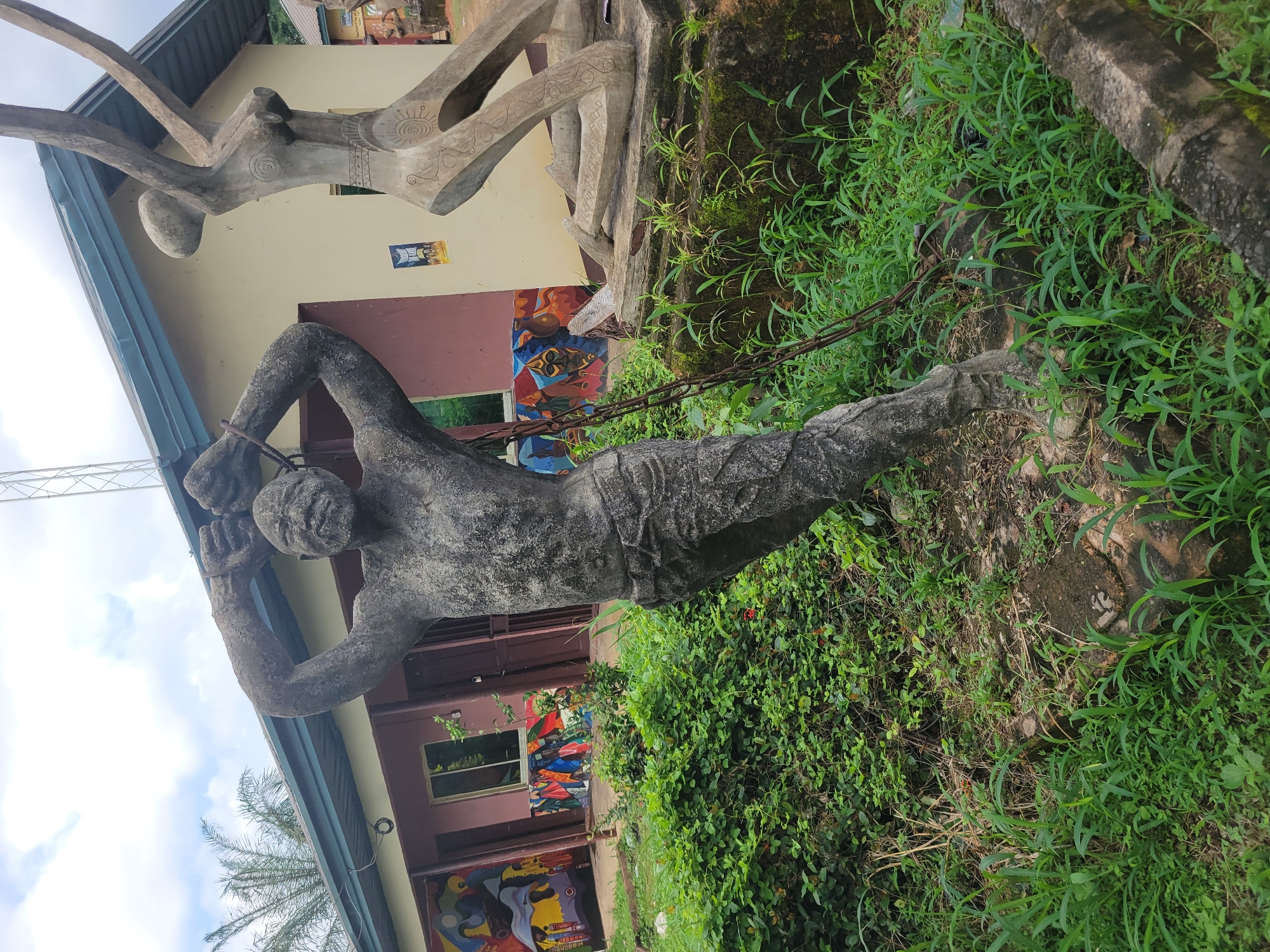 The image depicts an outdoor art installation in Egbagu, Nigeria. In the foreground, a weathered, grey statue of a man stands with his hands raised and bound by chains around his wrists. The statue is detailed, showing musculature and a strained expression. To the right, a second, abstract statue of a figure in a kneeling or rising pose is visible. Both sculptures are situated amidst lush green vegetation, including tall grass and bushes. Behind the statues, a building with a light yellow facade and a dark blue roof is visible. The building has several windows and doors, and the walls are adorned with colorful murals featuring abstract designs and figures. The sky above is overcast. The scene suggests a public art space or a cultural center.
