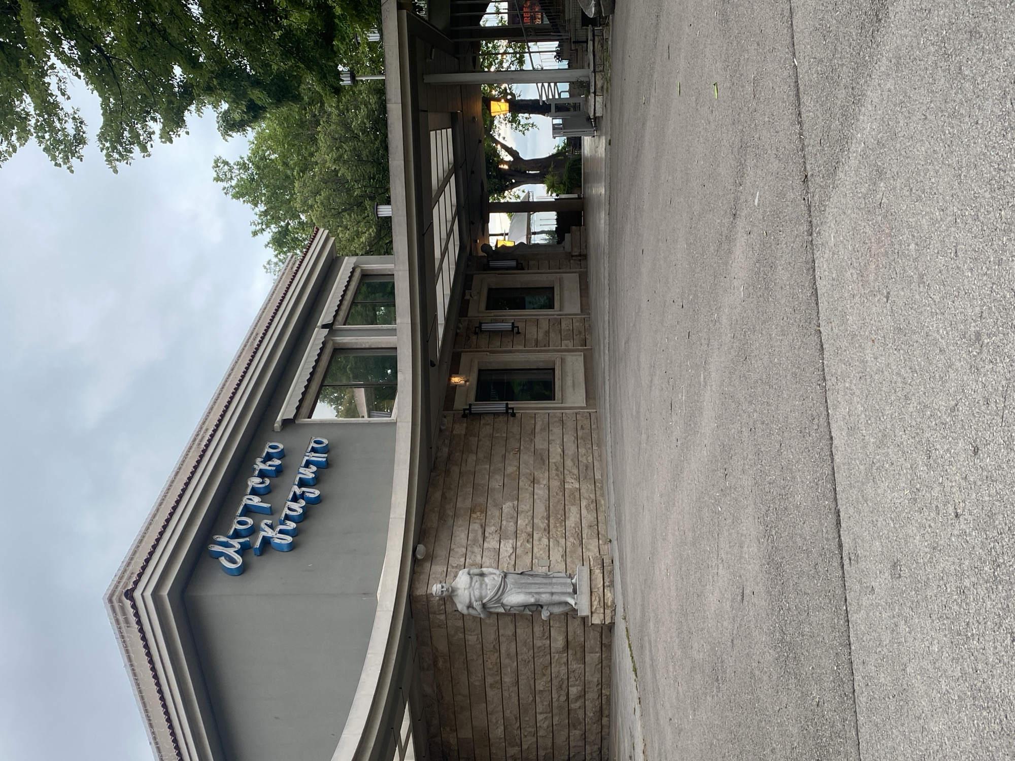 The image displays the exterior of a building, likely a public or recreational establishment, under an overcast sky during daytime. The building, identified as "Морско казино" (Morsko Kazino, or "Sea Casino") by the prominent blue Cyrillic text on its gray facade, is situated in Varna, Bulgaria.
The architecture features a structured roofline with multiple layers and windows along the upper floor. A section of the building below the signage is clad in light brown, vertically paneled material. Standing against this textured wall is a white, classical-style statue of a male figure, depicted in flowing drapery and mounted on a stone pedestal.
An outdoor covered area, supported by concrete pillars, extends from the building's entrance, featuring hanging lights. Beyond this structure, green foliage from trees is visible, suggesting a natural backdrop or parkland. The foreground and immediate right of the building are paved with gray concrete or asphalt. No people or specific activities are visible, presenting a static view of the building and its immediate environment.