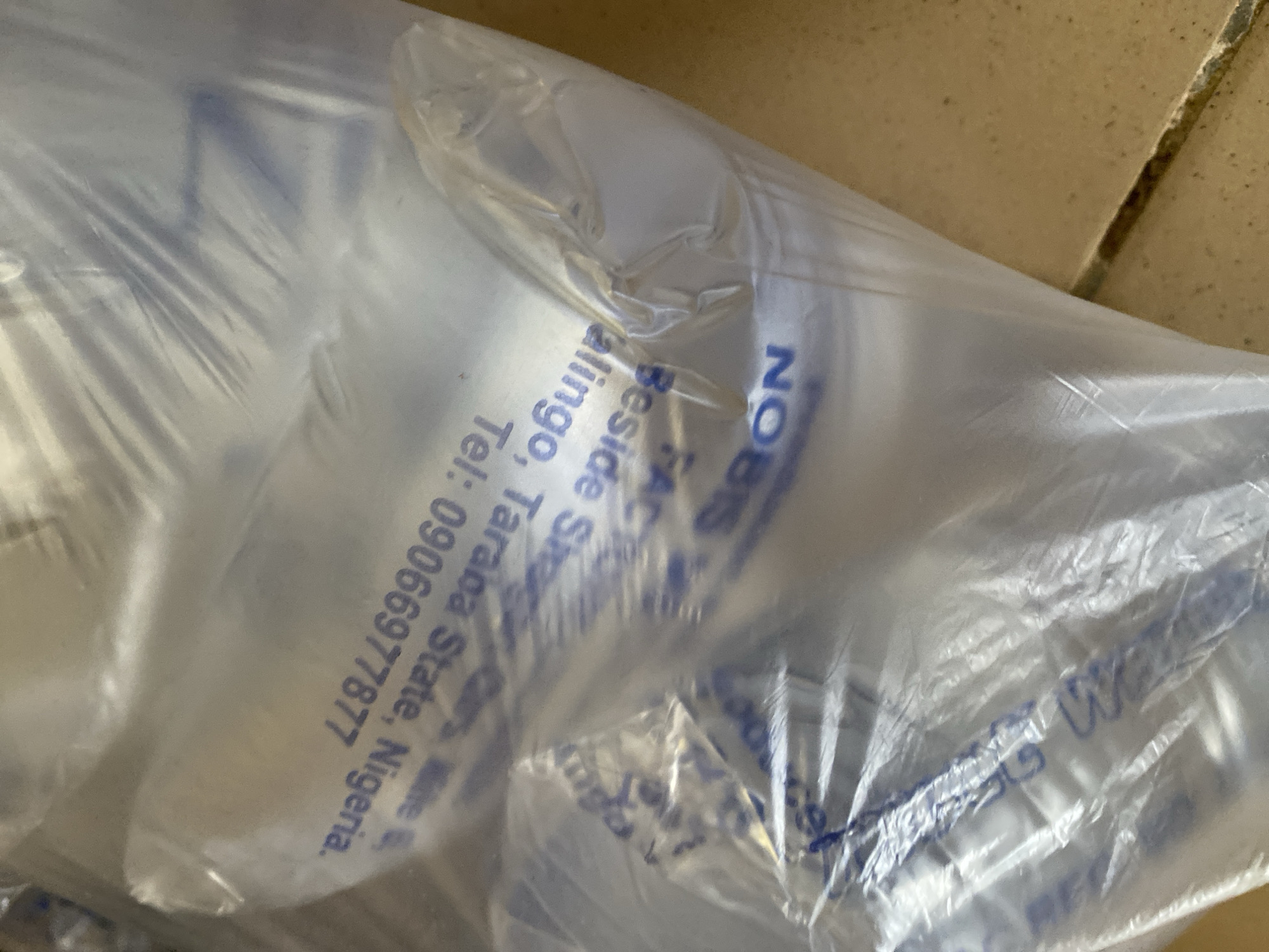 This is a close-up, angled shot of a translucent plastic bag containing cylindrical objects. The bag is crinkled and shows signs of wear, with folds and creases obscuring some of the details. The background appears to be a textured, light brown surface, possibly a floor or wall with tiles.
Visible text printed in blue ink on the bag provides information about the contents or their origin. The text reads: "NOBIS Beside ShCars Mie FACT alingo, Tarana State, Nigeria. Tel: 09066977877". The text is partially obscured by the plastic and the angle of the shot, but key elements like "NOBIS," "Tarana State, Nigeria," and a phone number are discernible. The overall impression is that of packaging for some form of product, possibly industrial or retail, with contact and location details provided. The lighting suggests it might be indoors or under artificial light, as there are no strong shadows or indications of natural light sources. No people or specific activities are depicted.