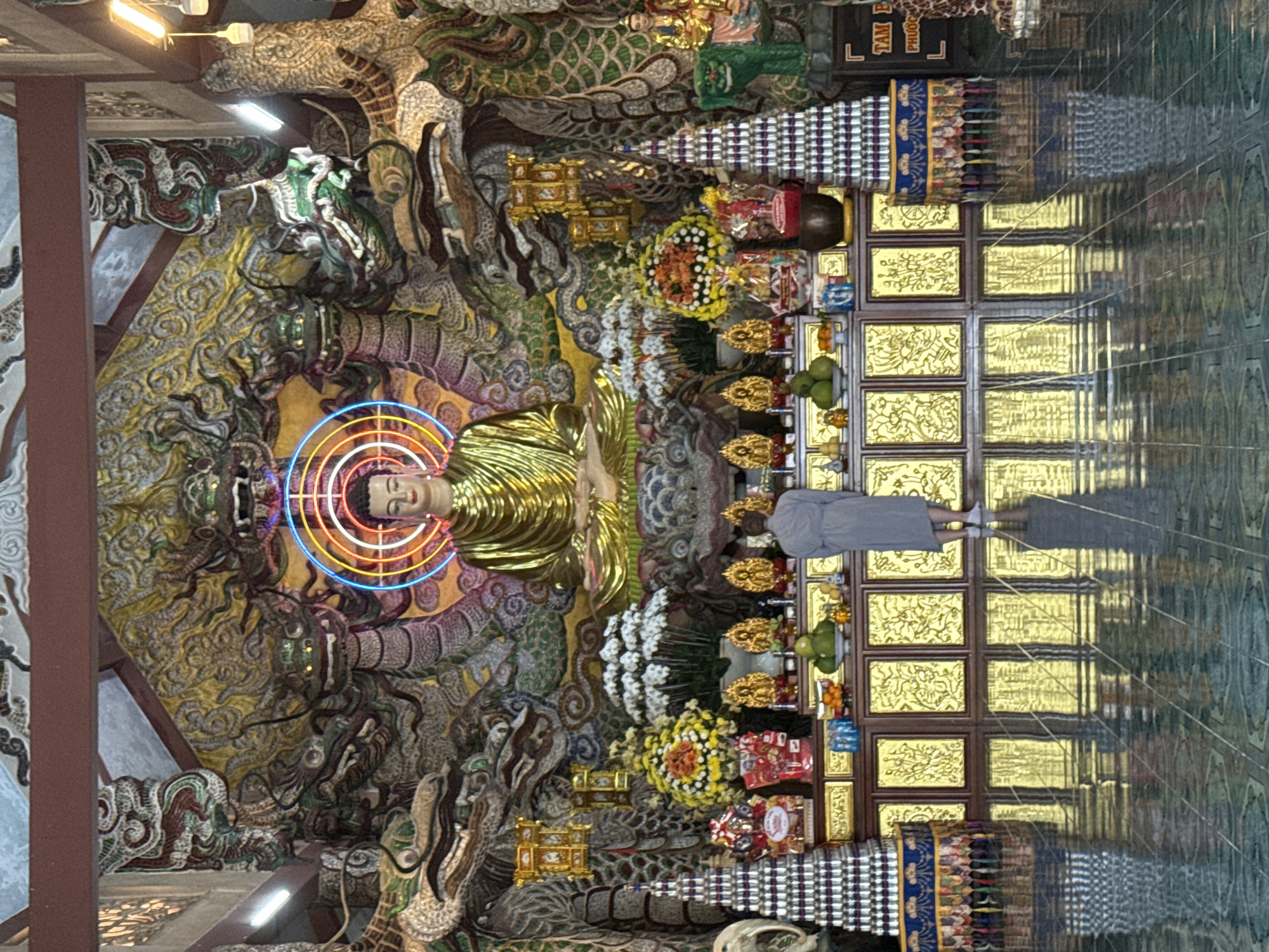 A person wearing a grey robe stands facing a golden Buddha statue in a temple in Lâm Viên - Đà Lạt, Vietnam. The Buddha is seated with a neon halo and is flanked by elaborate dragon carvings and floral arrangements. The floor is highly reflective, mirroring the temple's ornate decorations.