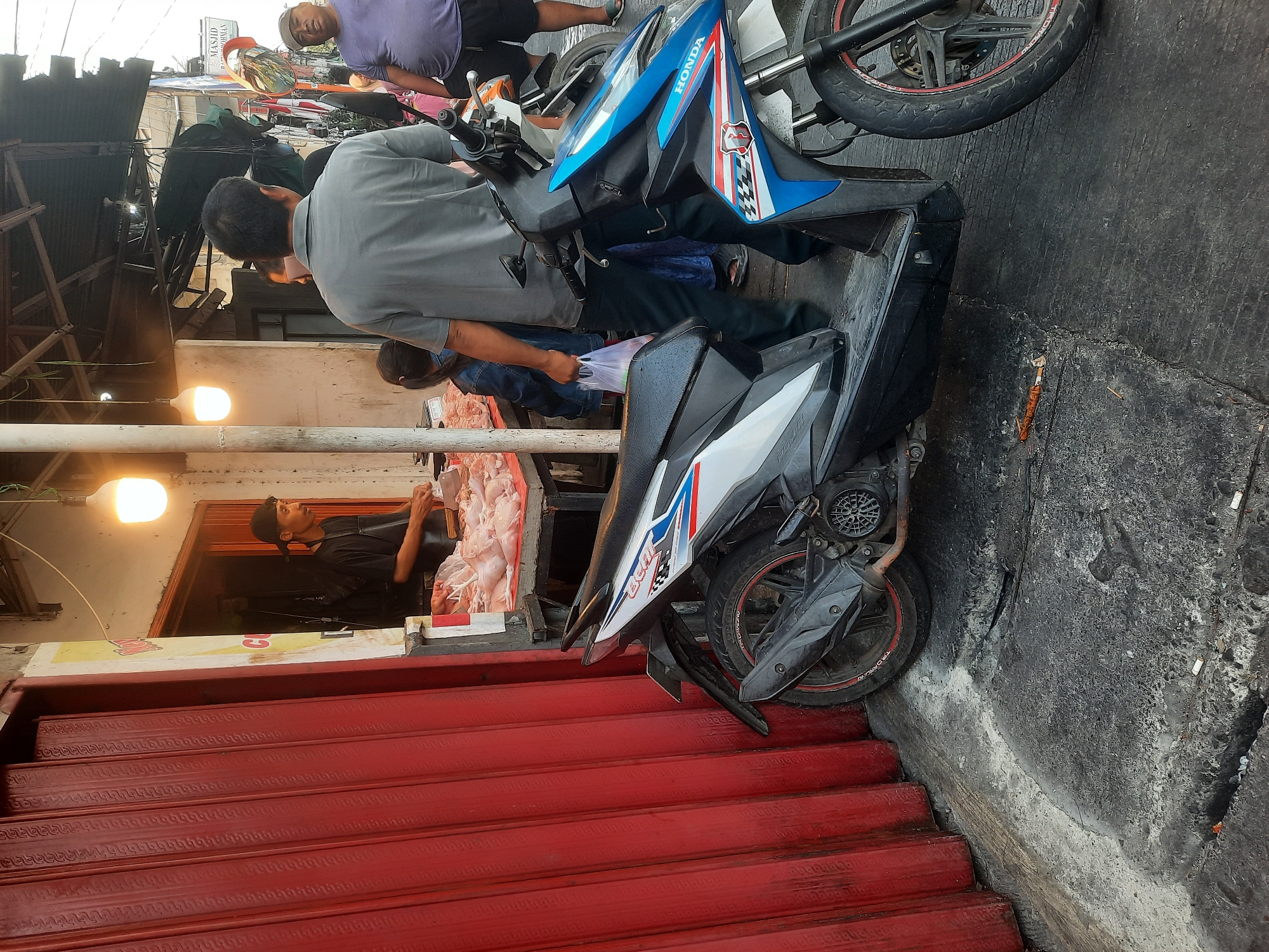 In North Jakarta, Indonesia, a butcher stands at his stall, preparing raw chickens. A red metal roll-up door is on the left. Two motorcycles are parked on the street in front of the stall. One motorcycle is white and black with red and blue accents, and the other is blue and white with red accents. A few people are standing around the stall, including a man in a grey shirt and dark pants, a woman holding a plastic bag, and a man in a purple shirt and shorts. The scene appears to be a street market.