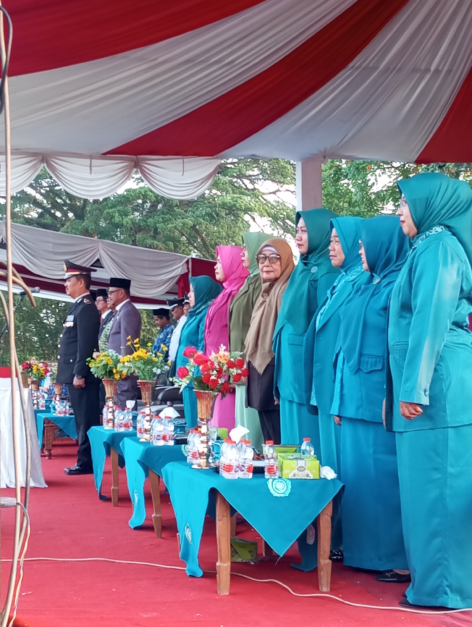 The image depicts an outdoor gathering on a red carpet, likely a formal event, under a large tent with red and white stripes. In the foreground, several tables draped in teal fabric are set up, adorned with bouquets of flowers, bottles of water, and other small items. A group of people are standing in a line, facing to the right. The individuals on the left are men in formal attire, including a man in a military-style uniform and another in a suit. To their right, a line of women is present, all wearing teal colored headscarves and matching outfits. They are positioned in a formal line, and their gaze is directed towards the right side of the frame. The background shows trees and foliage, suggesting the event is taking place in an open area, possibly a park or a designated event space. The presence of a microphone stand on the far left indicates a possible ceremony or speech. The overall atmosphere appears to be one of order and formality. The event is taking place in Tanjung Gading, Indonesia.