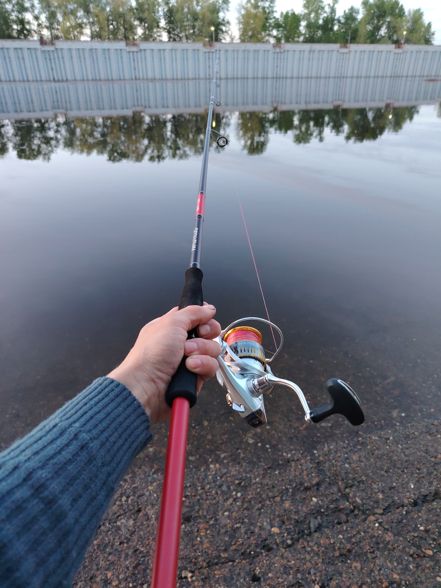 A first-person perspective image shows a person fishing in the city of Chaikovsky, Russia. The person is holding a fishing rod and reel in their right hand, with the line extended out into the water. The fishing rod is red and black with the word "SQUADRON" printed on it. The reel is silver and red. The person is wearing a blue sweater. The background shows a body of water, likely the Kama River, with a concrete wall along the shore and trees on the other side. The water reflects the wall and trees. The ground near the person is made of small rocks and pebbles.