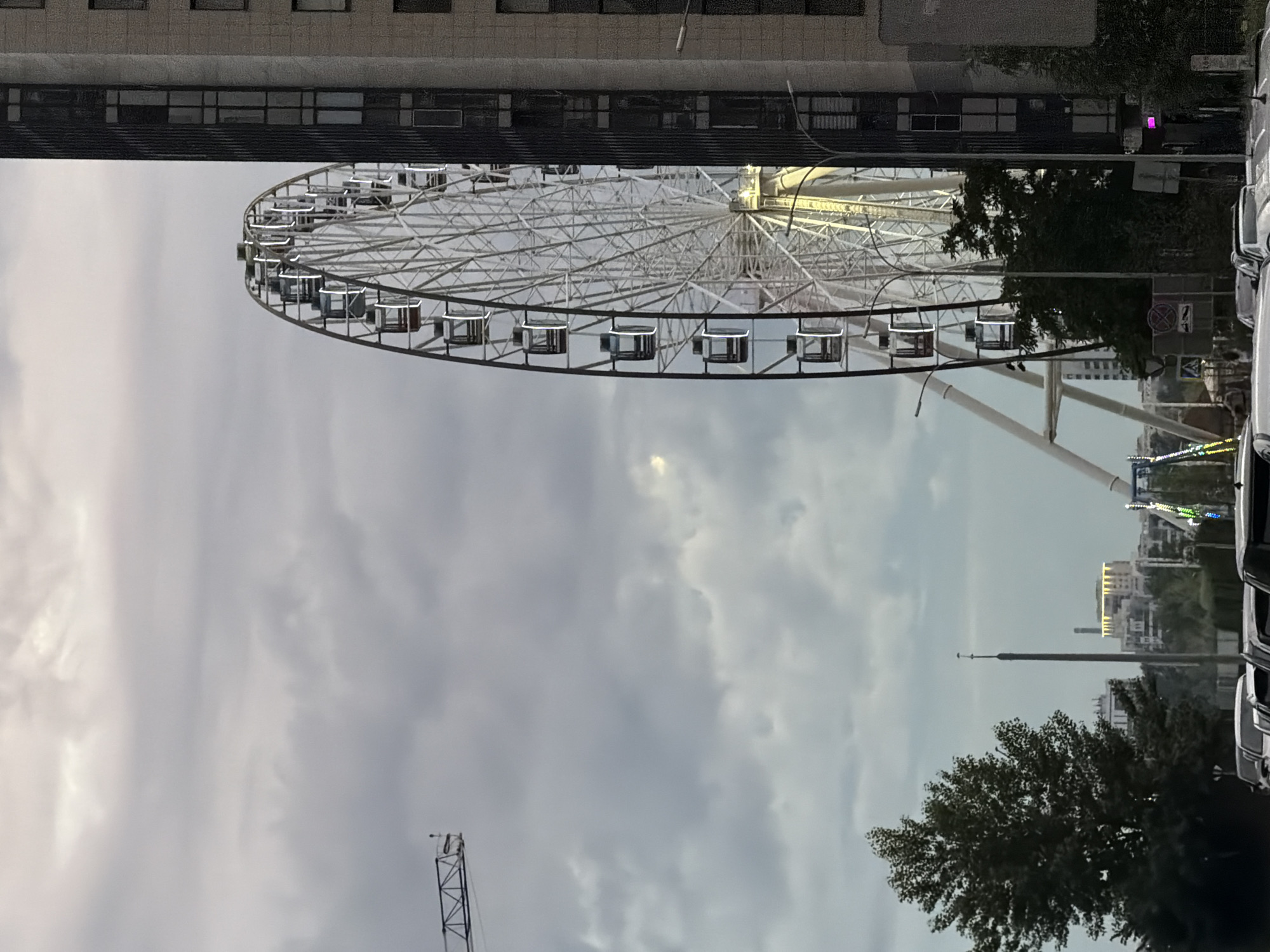 This is a photograph of a Ferris wheel in Kazan, Russia. The Ferris wheel is large and white, with many gondolas. It is set against a cloudy sky. In the background, there are buildings and trees. The photograph appears to be taken during the day, and the weather is overcast. The Ferris wheel is not in motion, and there are no people visible. The scene evokes a sense of calm and perhaps a touch of melancholy due to the cloudy weather. The image is a bit abstract, focusing on the structure of the Ferris wheel and the atmospheric conditions. The overall impression is one of a quiet day in a city park or amusement area.