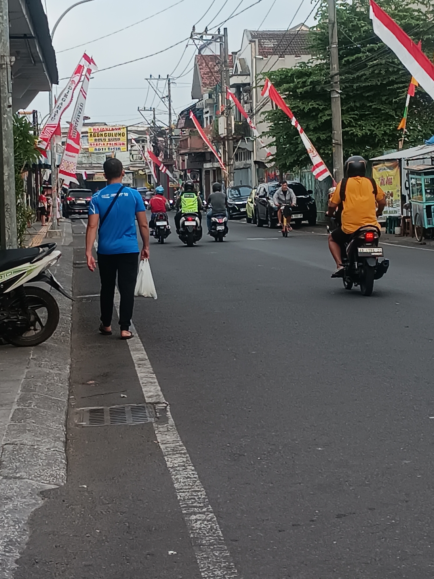The image depicts a street scene in Yogyakarta, Indonesia, likely during a festive period given the prevalence of Indonesian flags. The street is paved, with a sidewalk on the left and a gutter visible. A man in a blue shirt and black pants walks away from the camera on the sidewalk, carrying a white plastic bag. Several people are on motorcycles in the street, including a person in a yellow shirt on a motorcycle in the foreground. There are buildings lining the street, some appearing to be shops or residences. Overhead, power lines and more flags are visible. The sky is overcast. The scene suggests normal street activity with a celebratory atmosphere.