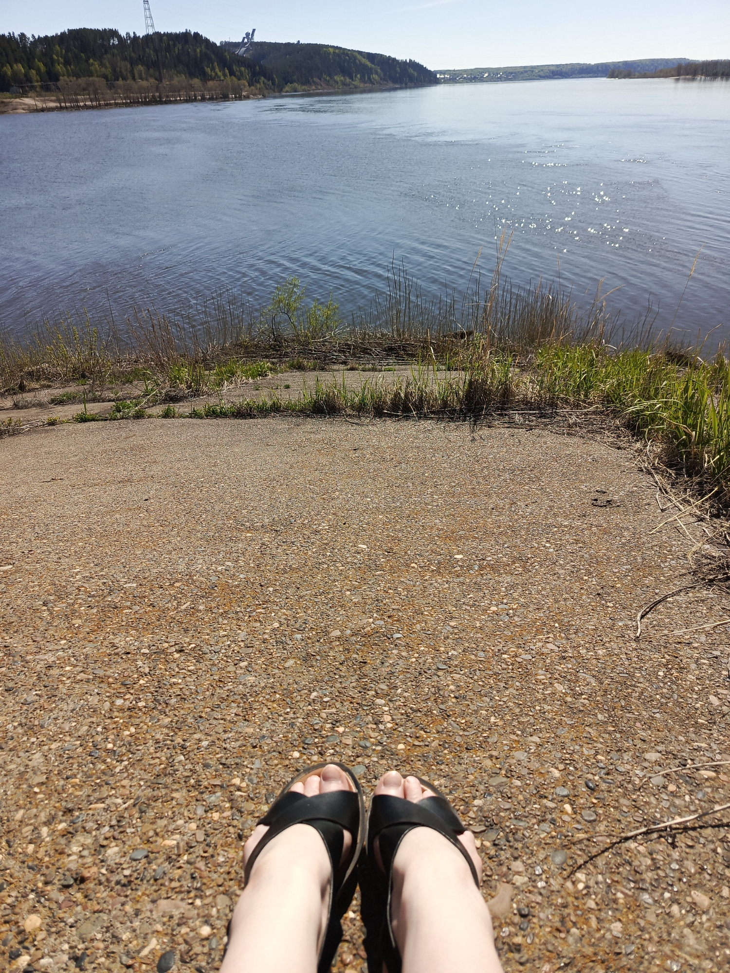A point-of-view, full-frame shot from ground level shows a person wearing black sandals on a concrete surface with small pebbles, looking out over a large body of water. The concrete surface slopes downward towards the water's edge, where vegetation grows. The water surface has ripples and sparkles in the sunlight. In the background, a wooded shoreline and a distant bridge or structure are visible. The scene is located in the city of Chaikovsky, Russia.
