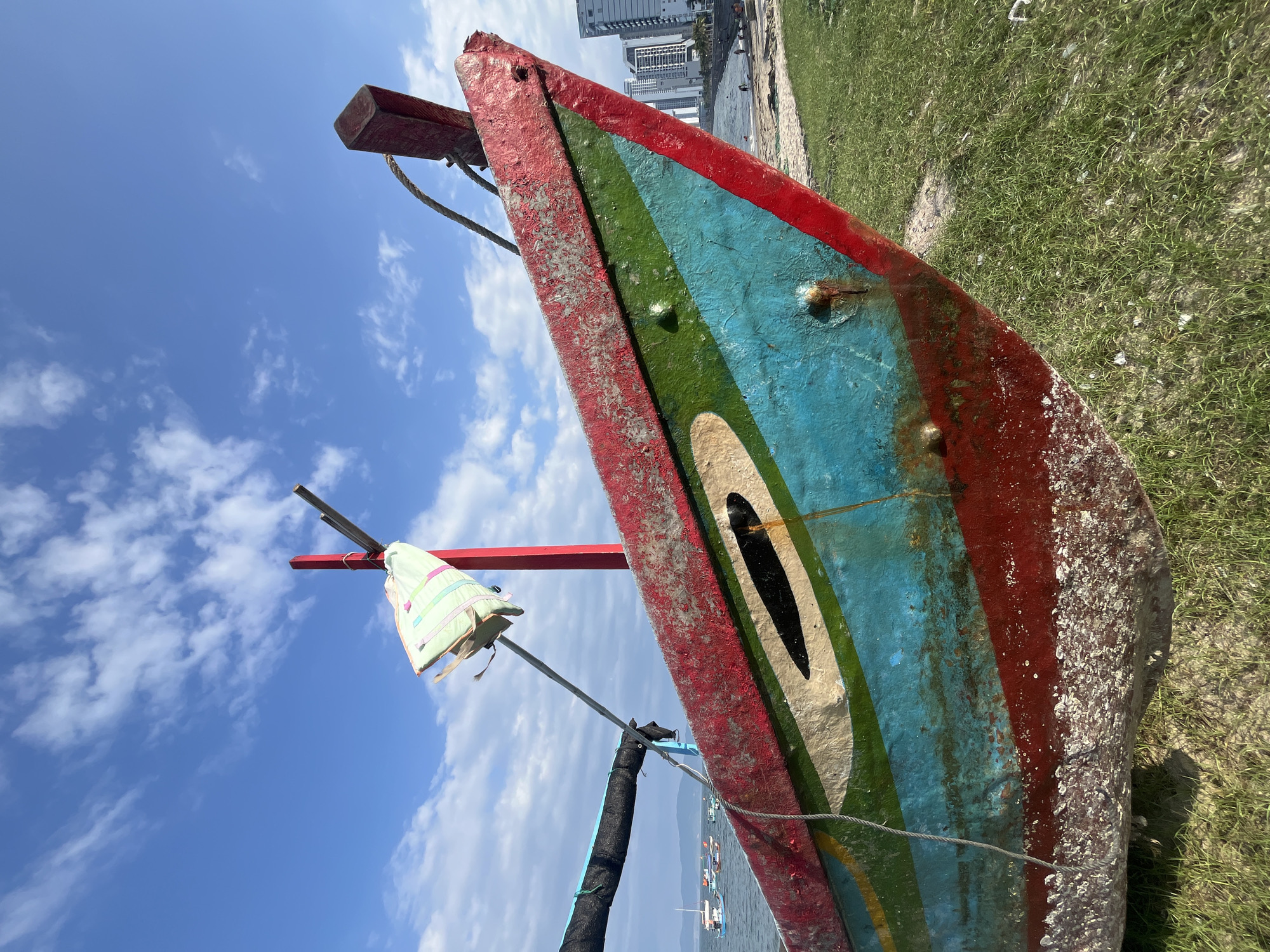 A weathered fishing boat, painted in worn bands of red, green, light blue, and off-white with black accents, occupies the foreground, angled with its bow directed towards the viewer and slightly right. The boat's lower hull is heavily encrusted with barnacles and marine growth. A red vertical pole rises from the boat, displaying a striped light green and pink fabric item, likely a life vest. A gray rope runs from the bow upwards and left. On the boat's left side, a dark material is visible wrapped around a horizontal boom. The boat is partially beached on sparse green grass and sand. In the mid-ground left, a calm blue sea features several other fishing vessels and distant land under a partly cloudy sky. To the mid-ground right, a sandy beach with scattered individuals leads to a modern city skyline dominated by multiple high-rise buildings. The sky above is clear blue with scattered white cumulus clouds, indicating bright daylight. The scene is located in Bac Nha Trang Ward, Vietnam, displaying a convergence of traditional maritime activity and urban coastal development.