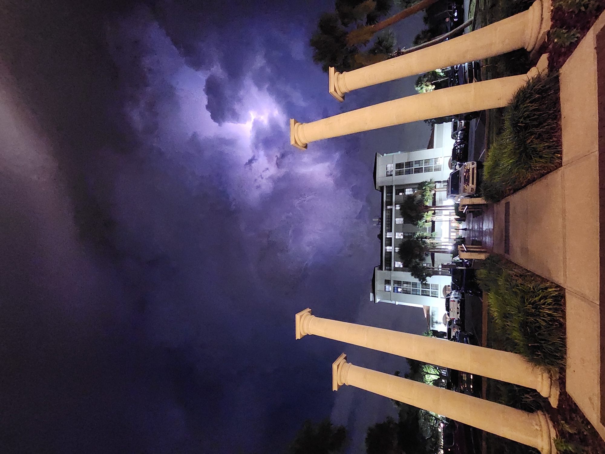 The image captures a dramatic nighttime scene in Altamonte Springs, United States of America, featuring a building with palm trees, parked cars, and a pathway lined with four large, cream-colored columns. A significant lightning strike illuminates the dark, cloudy sky. The building is lit from within, and its windows are visible. The foreground is dominated by the pathway and the bases of the columns, with manicured grass and landscaping. The perspective is from ground level, looking towards the building.