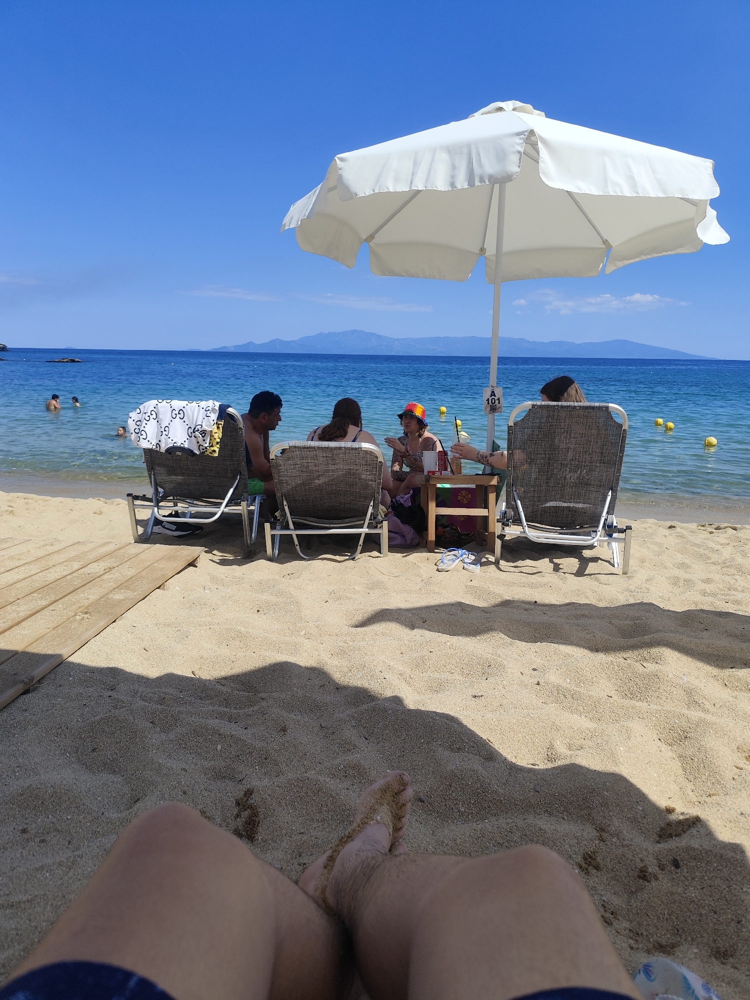 The image depicts a scenic beach scene in Kavala, Greece, with a clear blue sky and calm sea. The foreground shows a person's legs covered in sand, indicating they are lying on the beach. In the midground, a white umbrella provides shade for a group of people seated on lounge chairs. Two individuals, a man and a woman, are visible in the foreground, facing away from the camera and towards the sea. A small table sits between them, with drinks and what appear to be snacks. Further back, two more individuals are seated, one of whom is wearing a colorful hat and appears to be holding a drink. Several yellow buoys are visible in the water, and some people are swimming in the distance. The beach is sandy, with a wooden pathway leading towards the lounge chairs. The background features a distant coastline with mountains under a bright sky.