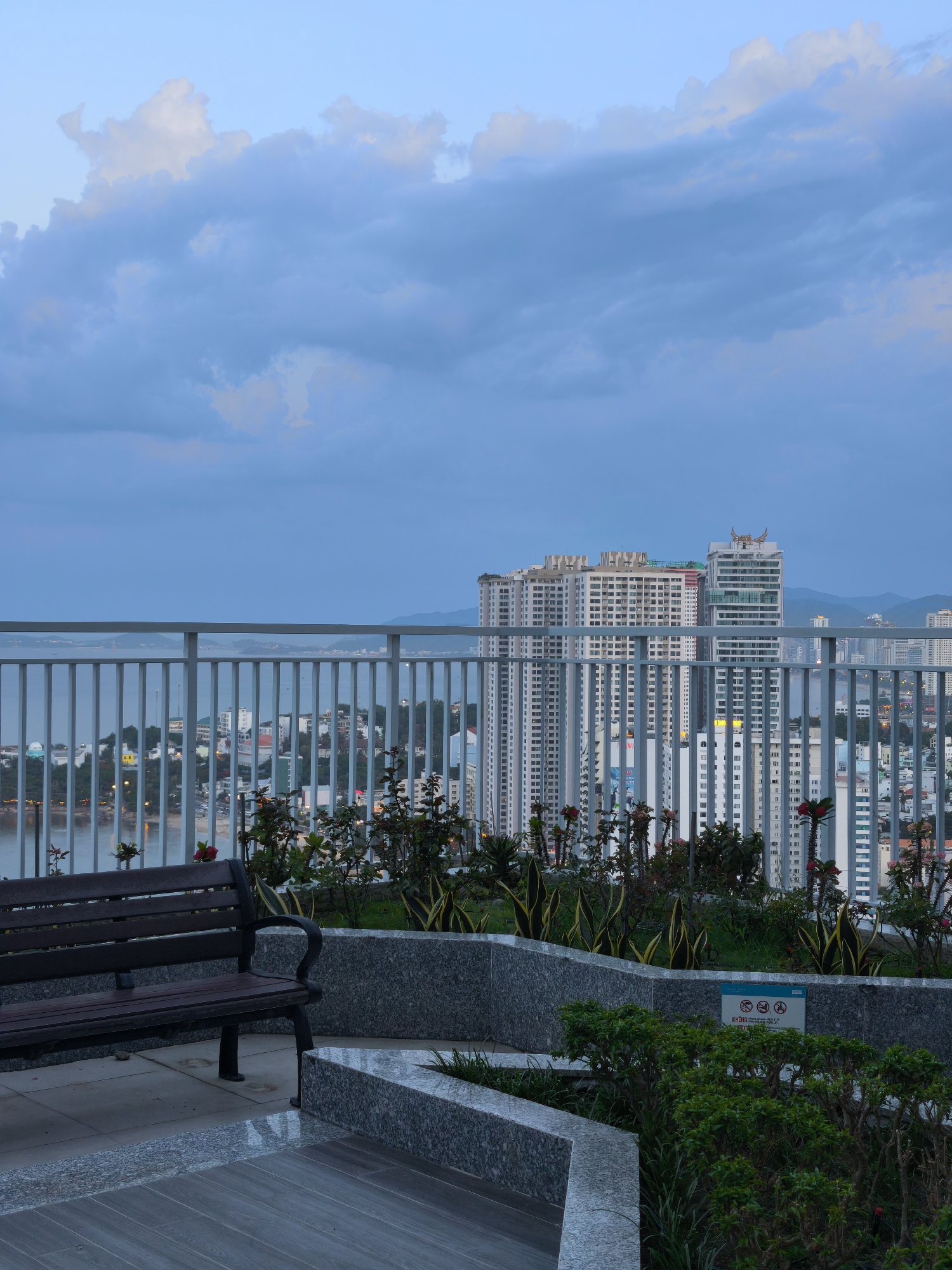 This media file depicts a serene cityscape from a high vantage point, likely a rooftop or balcony in Bac Nha Trang Ward, Vietnam. The scene is set during dusk, as indicated by the soft, diffused light and the twilight blue of the sky, which is adorned with dramatic, billowy clouds.
In the foreground, a dark wooden park bench sits on a tiled patio, offering a place for contemplation. Adjacent to the bench is a planter box filled with lush green plants and flowering bushes, some with hints of red blossoms. A metal railing with vertical bars separates the foreground from the view of the city and the sea beyond.
The middle ground reveals a sprawling urban landscape dominated by numerous tall, modern apartment buildings that stretch towards the horizon. Their windows, reflecting the fading light, create a textured pattern against the sky. A body of water, likely the sea or a bay, is visible to the left, with boats and distant lights hinting at activity. In the far distance, rolling hills or mountains are faintly visible, shrouded in the evening haze.
A small sign is affixed to a low wall in the foreground, featuring several pictograms and text, though the specific details of the text are not clearly legible in the main image. The overall mood is one of tranquility and calm, inviting viewers to soak in the expansive vista as day transitions into night.