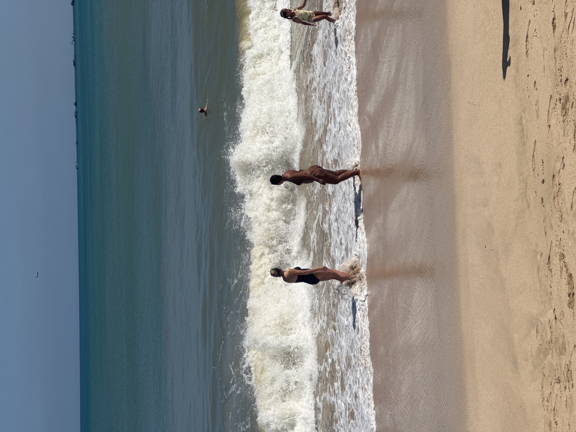 In the city of Kuta Selatan, Indonesia, an ocean beach scene unfolds under a clear, bright sky. The water, a mix of deep blue and turquoise, meets a sandy shore where several individuals are present.
In the foreground, wet sand shows subtle patterns from receding waves. Further out, a large wave is breaking, its white foam creating a dynamic line across the frame. Two adult women are standing in the shallow water near the breaking wave. The woman on the left, facing away from the camera, wears a black swimsuit. The woman on the right, also facing away, is in a patterned bikini. A young girl, wearing a yellow swimsuit, stands in the surf to the right of the two women.
In the mid-ground, further out in the ocean, a person is swimming. In the background, the horizon is lined with several small boats, and even further in the distance, a tiny airplane is visible in the sky. The overall atmosphere is one of a typical beach day with recreational activities.