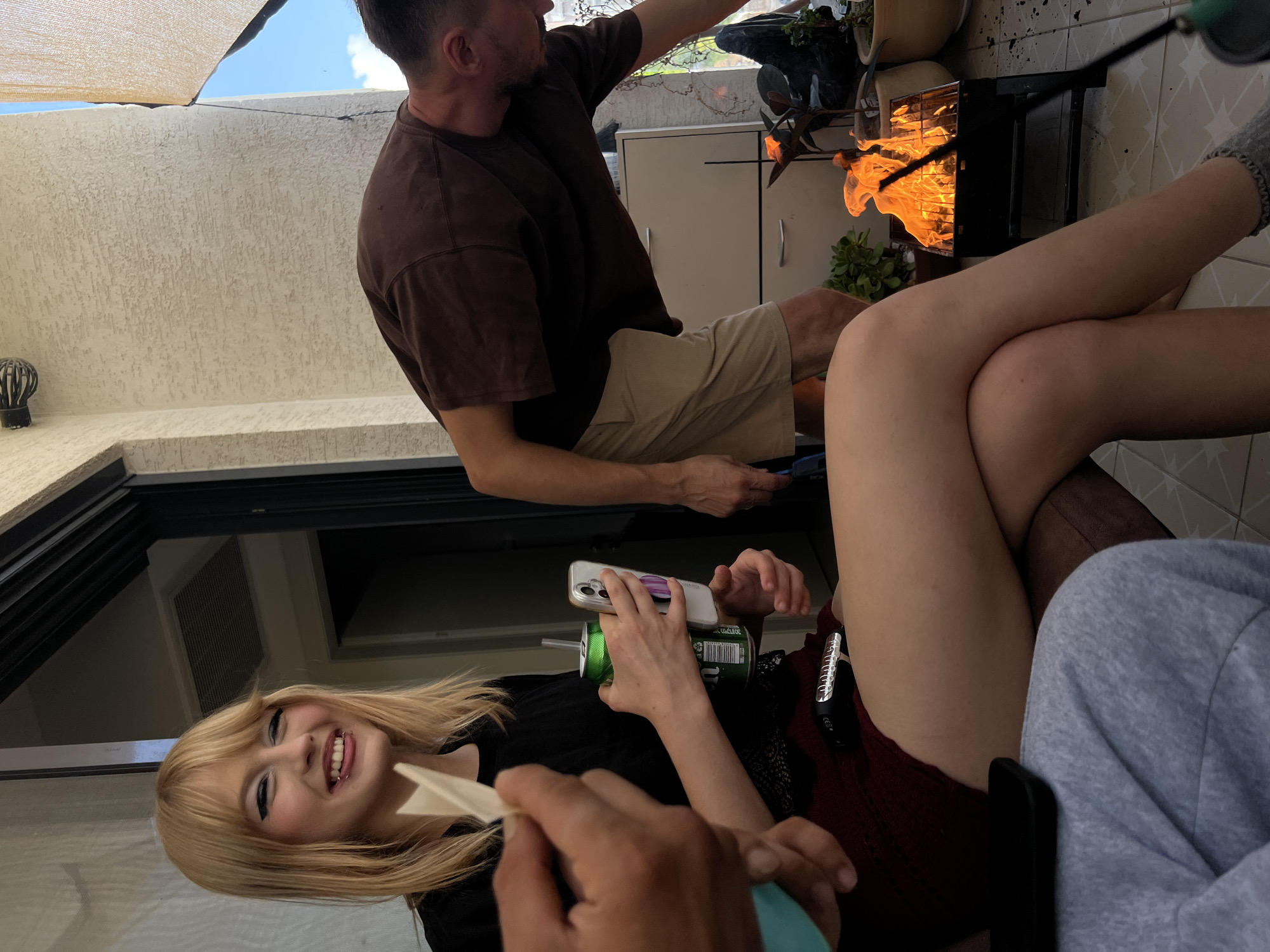 The image depicts an outdoor scene, likely a balcony or patio, where a small barbecue grill with visible flames is set up. A woman with blonde hair, wearing a black top and a dark skirt, is seated and holding a green can and a smartphone. She is smiling and has dramatic eye makeup. To her right, a man in a brown t-shirt and tan shorts is bending over the grill, seemingly tending to it. Another person's arm is visible in the foreground, holding what appears to be a rolled paper object. The environment suggests a casual gathering. The image was captured in Kiryat Bialik, Israel.