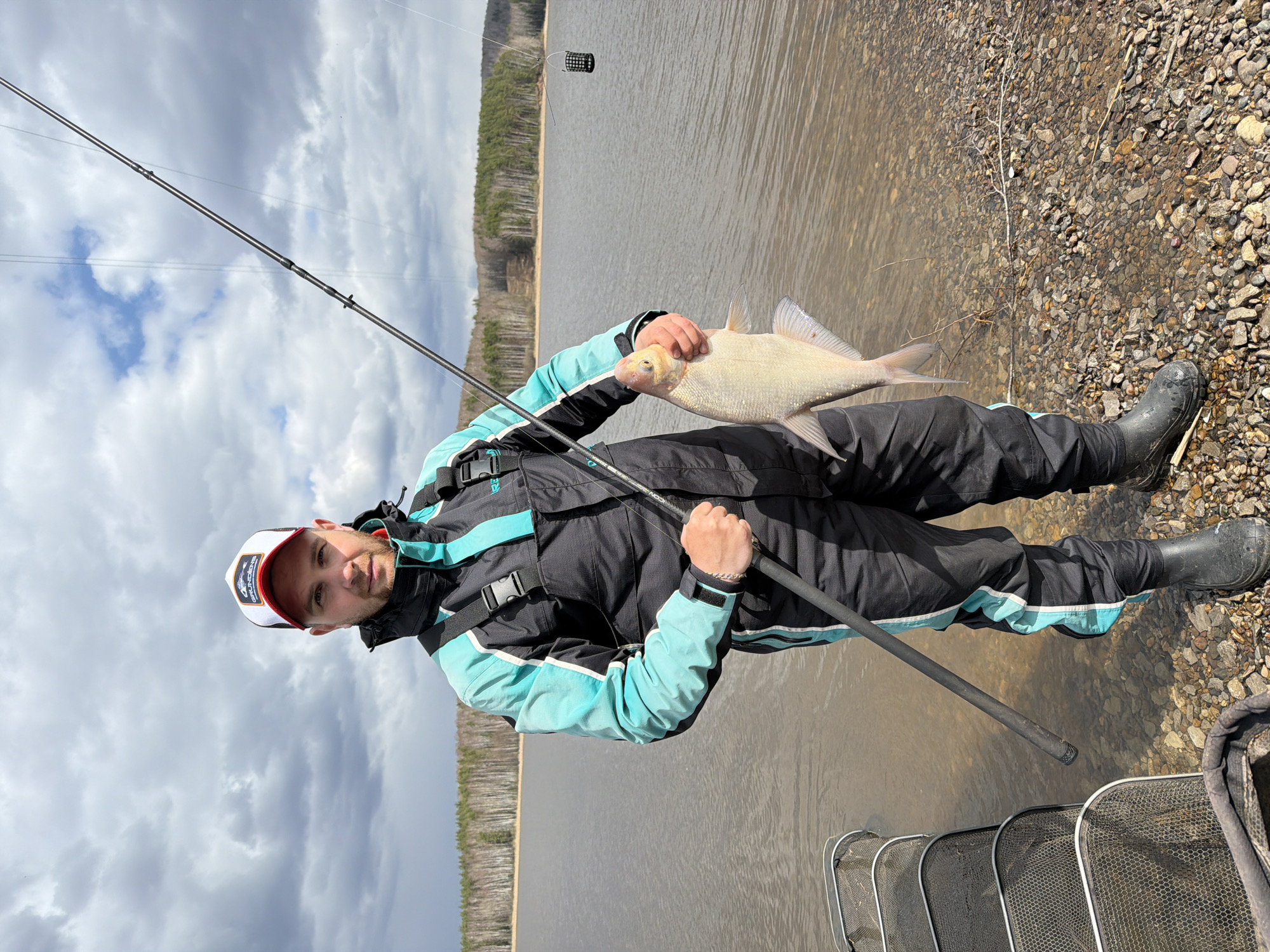 A man is captured in a full-length shot, standing on a rocky shoreline and holding a large, light-colored fish. He is engaged in fishing, with various pieces of fishing gear visible.
The subject is a man, likely in his 20s or 30s, with short facial hair, wearing a functional fishing suit consisting of a black and teal/light blue jacket and matching pants. He sports black rubber boots and a white baseball cap with a red brim, on which the brand "GRUNDÉNS" is partially visible. He holds a fishing rod, which has "ICON" visible on it, in his left hand while cradling the fish with both hands. His gaze is directed towards the camera with a neutral expression.
The fish he holds is substantial, with silvery-white scales. Near the water's edge, several net frames, possibly a keepnet or parts of a landing net, are laid out. A fishing feeder basket is suspended by a line a short distance from the shore in the water.
The setting features a large body of water, likely a lake or reservoir, with a treeline or forest visible on the distant shore. The immediate ground underfoot is composed of small stones and pebbles. The sky is largely overcast with white and grey clouds, indicating a cloudy daytime. The scene suggests an outdoor fishing activity in a natural, possibly cool, environment, consistent with locations like Chaikovsky, Russia.