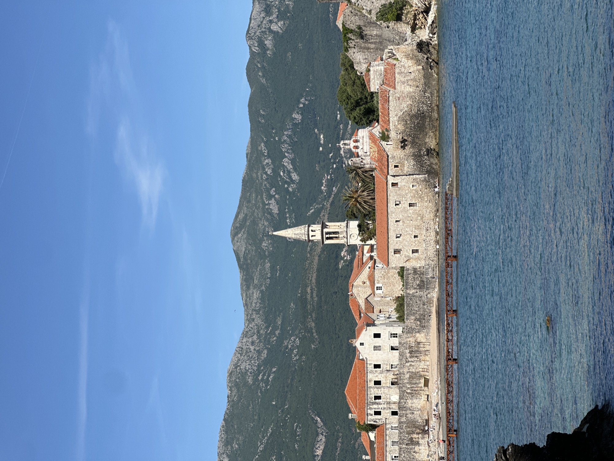 The image depicts the island of Sveti Stefan, a luxury resort near Budva, Montenegro. The island features stone buildings with red-tiled roofs clustered together, surrounded by defensive walls. A narrow isthmus connects the island to the mainland. The Adriatic Sea borders one side, while a steep, mountainous landscape rises on the opposite side. A blue sky with wispy clouds is visible above. The overall impression is a picturesque coastal scene with a blend of natural beauty and historic architecture.