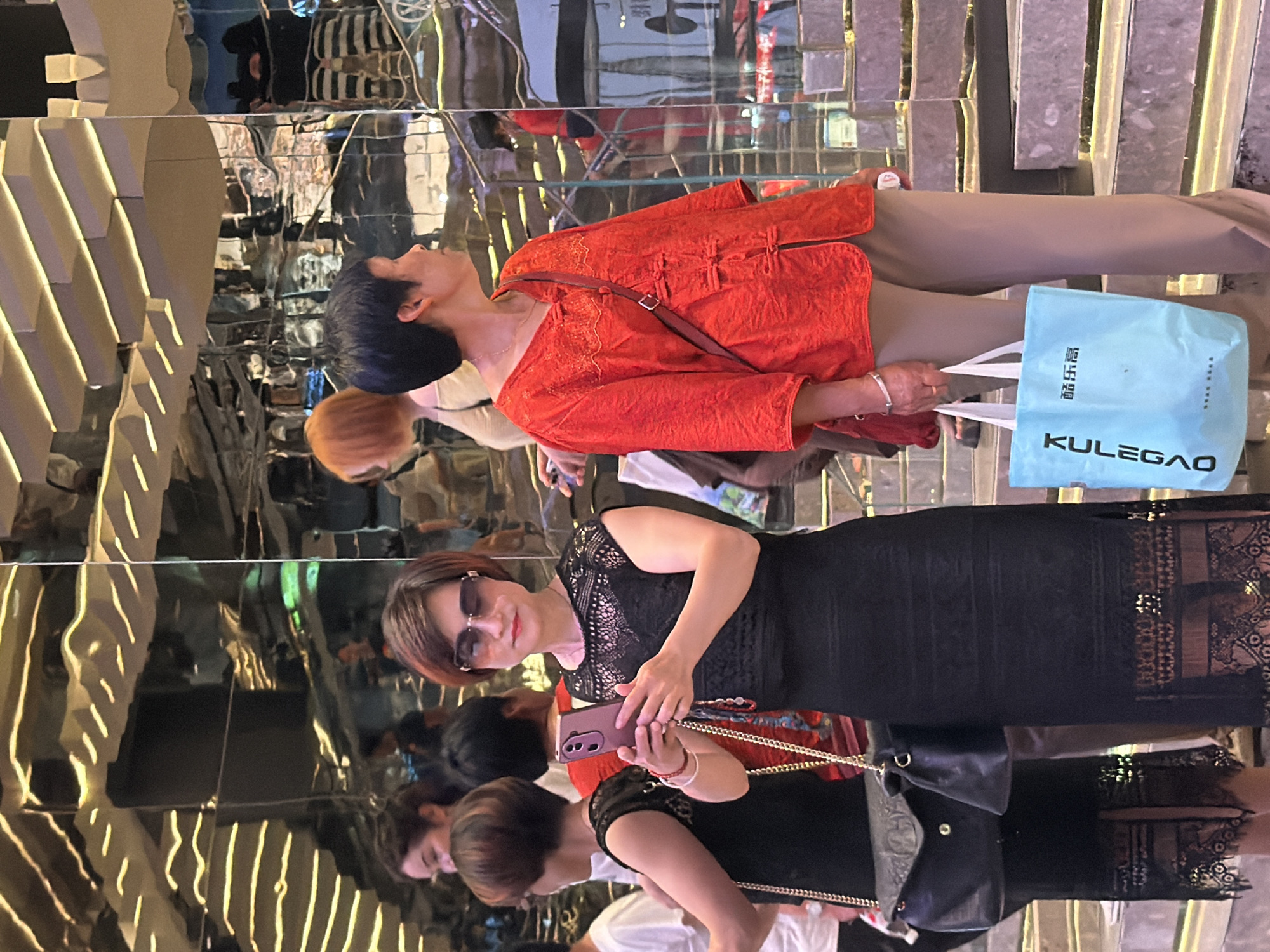 In Yuzhong District, China, two women stand near a mirrored staircase. The woman on the left, wearing a black lace dress and sunglasses, holds a pink smartphone. Her companion on the right, dressed in a red embroidered tunic and beige trousers, carries a light blue shopping bag with Chinese text and the brand "KULEGAO." The background features reflections of architectural elements and other individuals, suggesting a bustling commercial or public space.