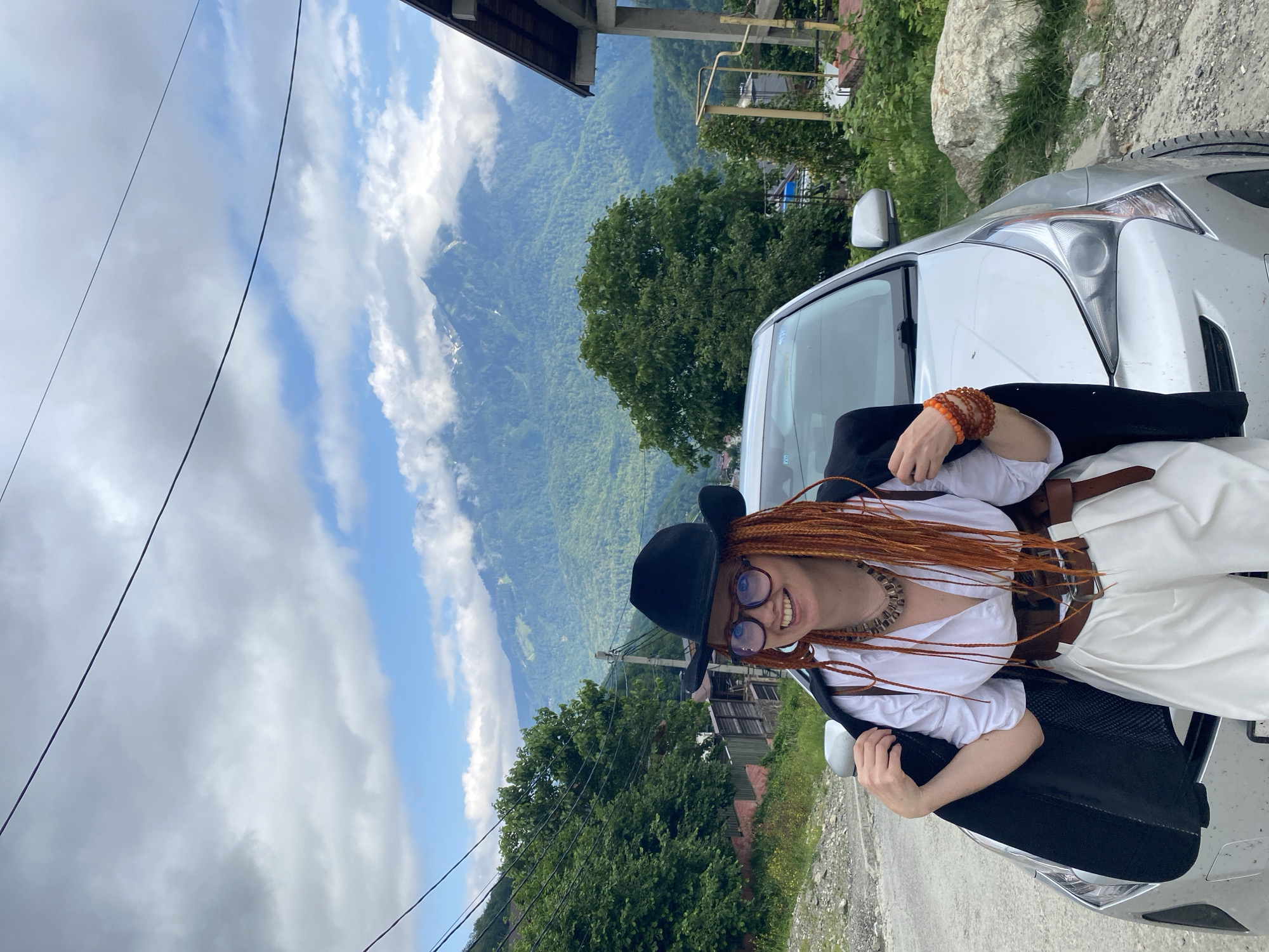 A woman with bright orange braided hair and glasses, wearing a black cowboy hat, a white collared shirt, a brown corset-style belt, and white wide-leg pants, stands next to a silver car. She is holding her black blazer open. The background features a lush green mountainside with scattered buildings and a cloudy sky. This scene is consistent with the natural landscape found in the vicinity of Krasnaya Polyana, Russia, a popular mountain resort area known for its scenic beauty and outdoor activities.