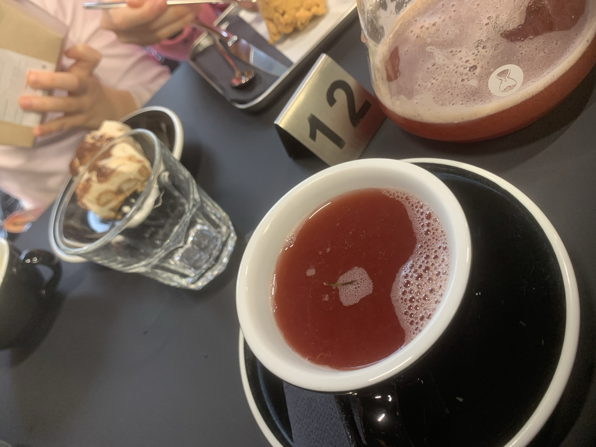 The image shows a table setting with various items: a clear glass containing a dessert with cream and light-colored chunks, a cup of red tea or juice in a white cup on a black saucer, a carafe containing a red beverage, a metal sign with the number "12", and a black tray holding a fried item with cutlery. A person in a pink shirt is visible on the left, holding a box or book. The scene appears to be in a cafe or restaurant setting. The location within the city of Izhevsk, Russia, cannot be determined from the image alone.