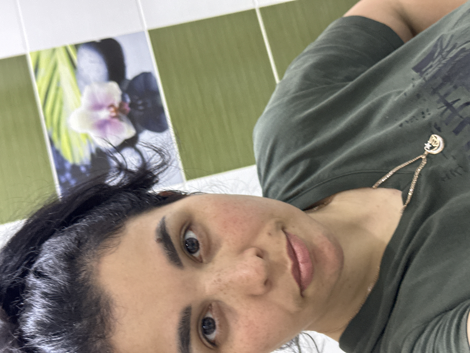 A selfie taken indoors, likely in a bathroom, of a woman with dark hair and fair skin. She is wearing a green t-shirt and a delicate gold necklace. The background shows green and white tiled walls, with a tile depicting a white and purple flower. The city and specific location are unknown.