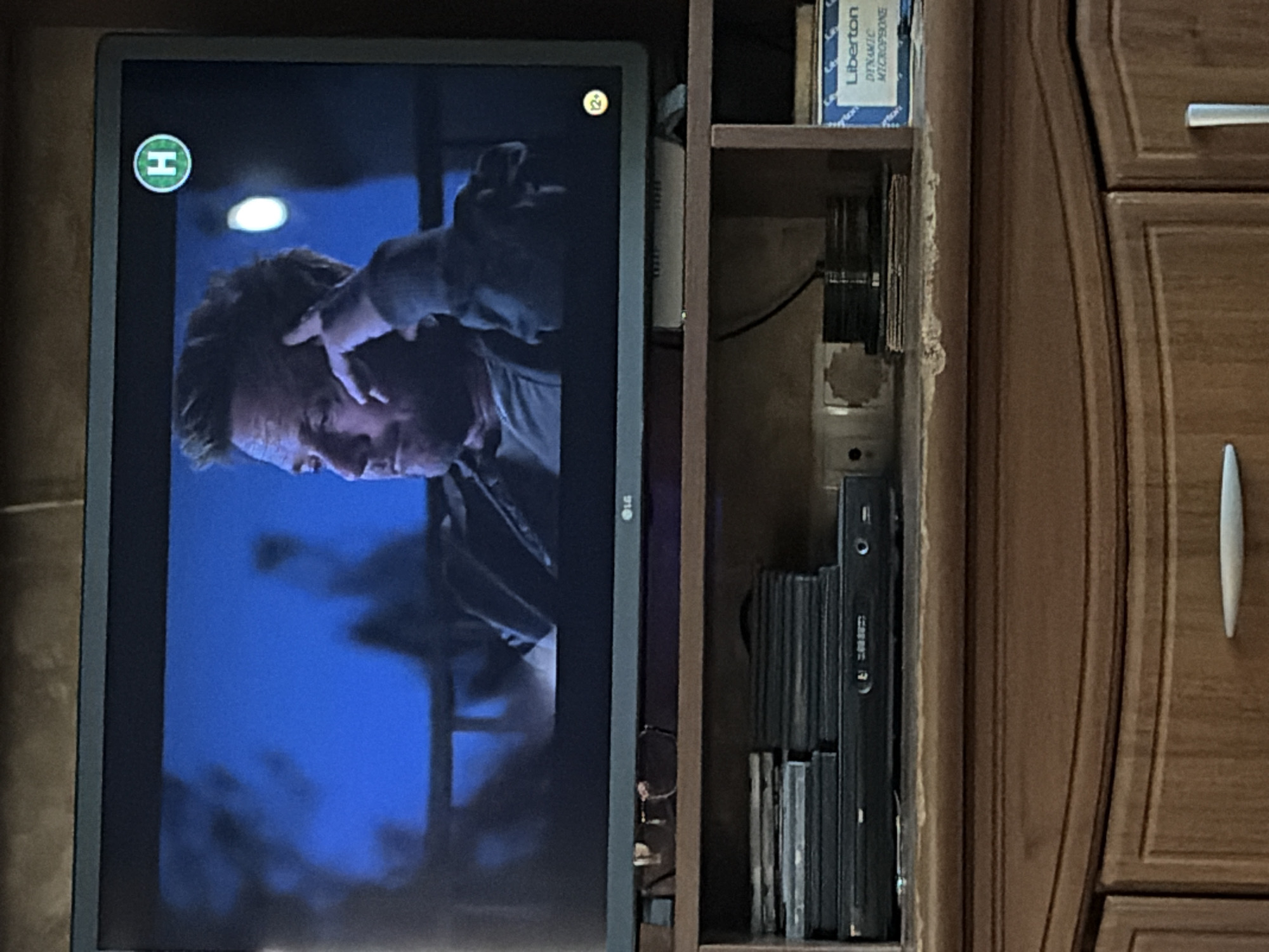 The image is a medium shot of a television screen displaying a man with his hand on his forehead. The screen shows a channel logo of a green circle with an "H" inside, located in the upper right corner. A yellow circle with "12+" inside is visible in the lower right corner of the screen, indicating a content rating. The television is placed on a wooden shelf unit, which also holds stacks of DVDs and a DVD player. A box labeled "Liberton DYNAMIC MICROPHONE" is also visible on the shelf. The background of the television screen is a dark blue, possibly depicting a night scene. The overall environment suggests an indoor setting, likely a living room or entertainment area. No specific city or location details are discernible from the image itself.