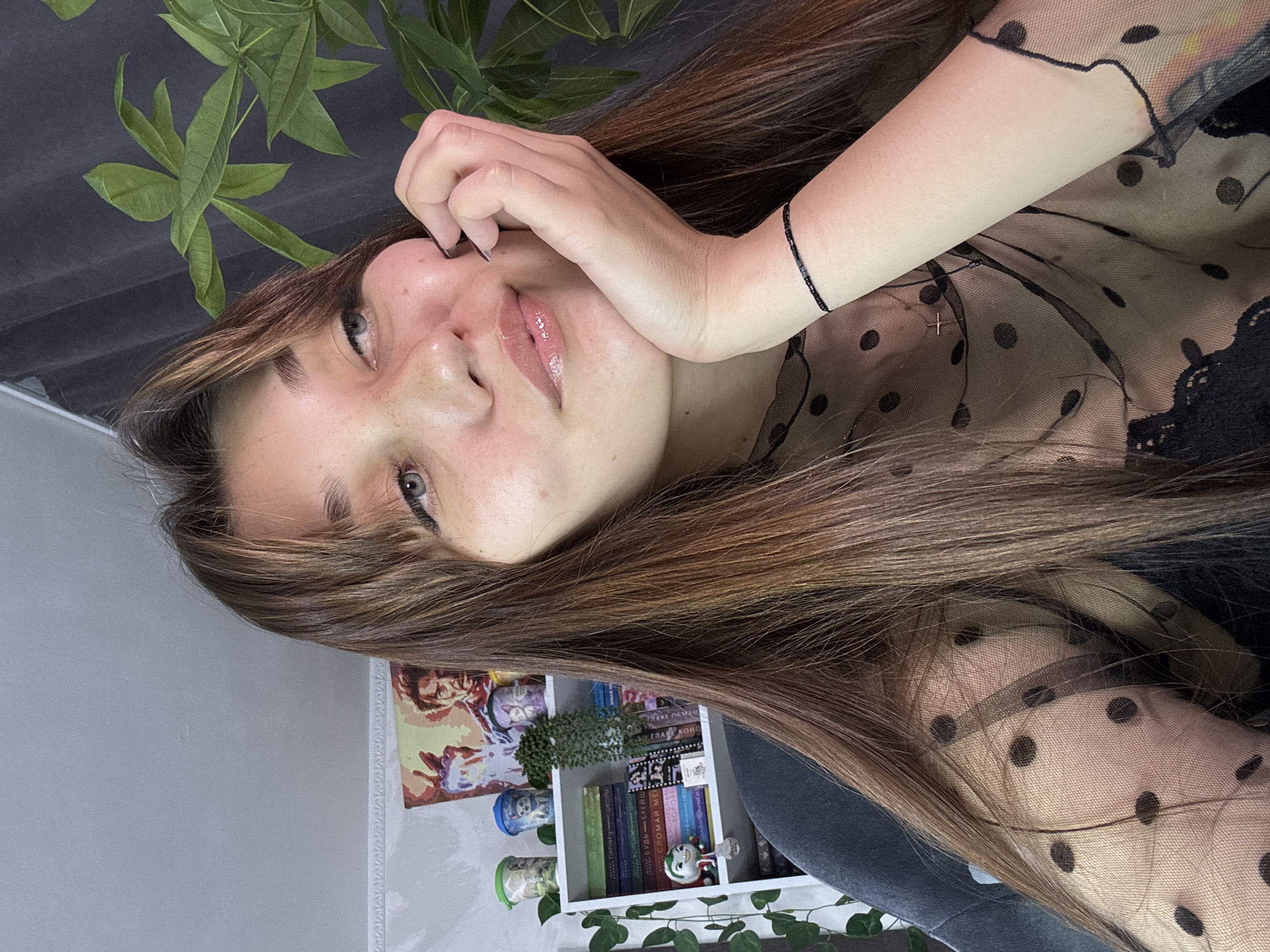 A young woman with long, light brown hair and bangs is captured in a close-up, medium shot, indoors. She is looking upwards and to her right, with her right hand supporting her chin. She wears a sheer black top with a polka dot pattern and a subtle cross necklace. Her skin has a dewy appearance, with visible freckles.
The background shows a white bookshelf filled with books, decorative cups, and a small figurine. To the left of the bookshelf, a poster featuring Tony Stark from Iron Man is visible. Lush green foliage from a plant is present on the right side of the frame, partially obscuring a dark curtain. The scene appears to be a home environment. There is no identifiable information to determine the location in Novokuznetsk, Russia.
