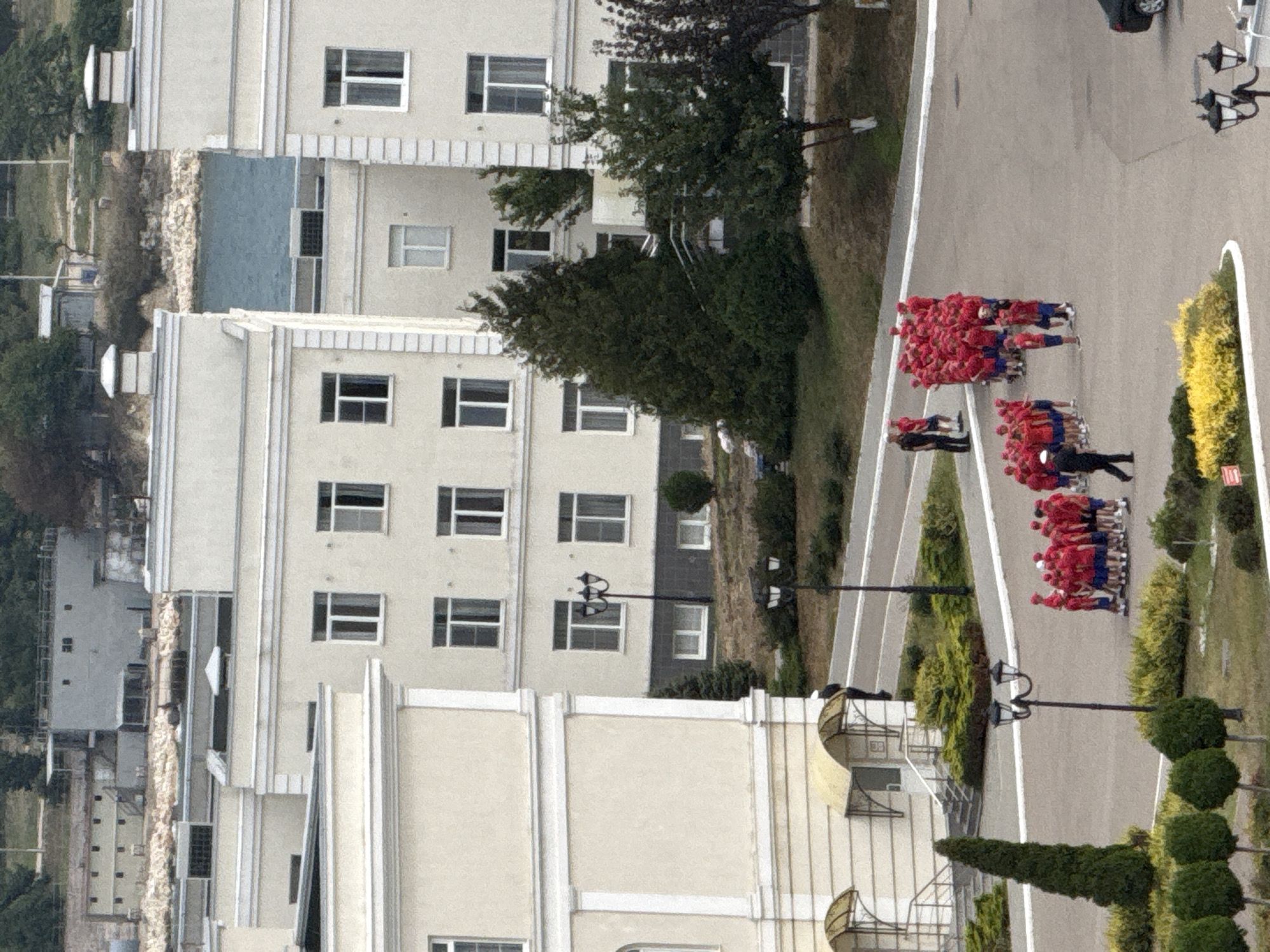 An aerial perspective captures a military or educational institution in Sevastopol, Ukraine, with multiple cream-colored buildings featuring rows of windows and decorative architectural elements. The buildings are set against a backdrop of green foliage and a body of water. In the foreground, a group of individuals, appearing to be students or cadets, are gathered outdoors, many wearing red shirts and blue shorts. Some are standing in formation while others appear to be observing or moving. One individual in a dark uniform stands out from the group, possibly an instructor or supervisor. Several trees and landscaped areas with bushes and yellow flowers are also visible on the grounds. The scene suggests an organized gathering or activity taking place at the institution.