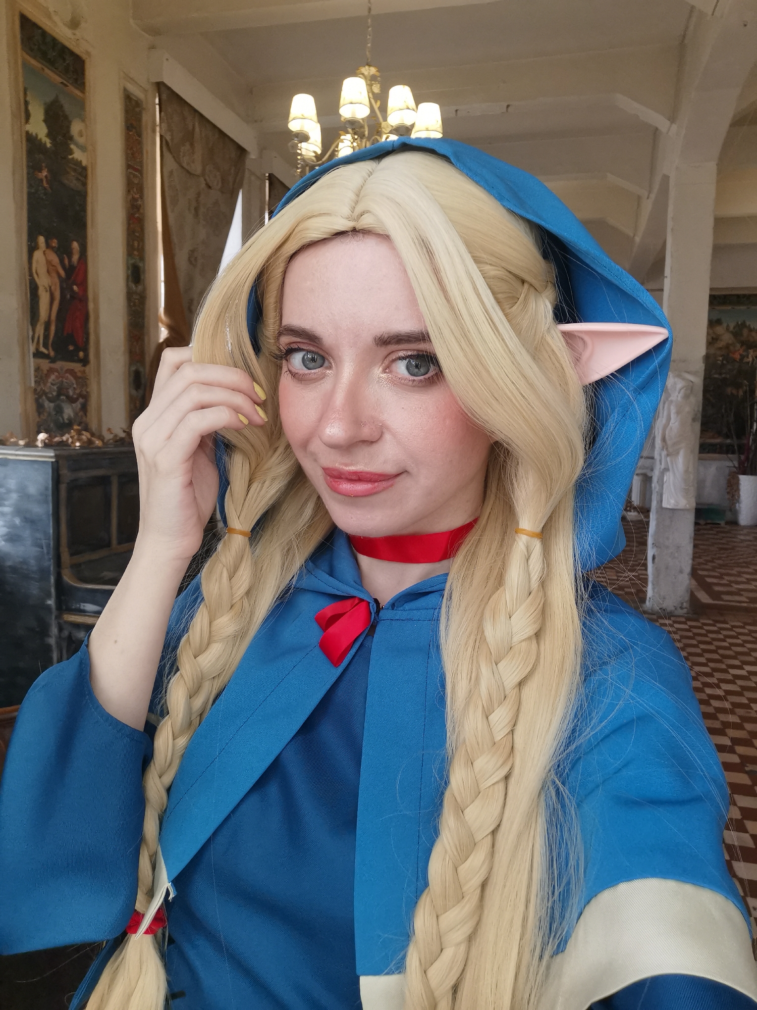 A woman, presumably located in Moscow, Russia given the architectural and decorative style of the building behind her, is pictured in a close-up selfie. She is wearing a blonde wig styled in two braids with rubber bands and an electric blue hooded garment with red ribbon details, suggesting a cosplay costume. The woman has fair skin, blue eyes, and a slight smile. In the background, there's a building with paintings on the walls, tall windows, and tiled floors, featuring an ornate chandelier. The presence of these interior details strongly implies a location in Russia, specifically Moscow, given its historical architectural influences and preservation of traditional decorative arts.