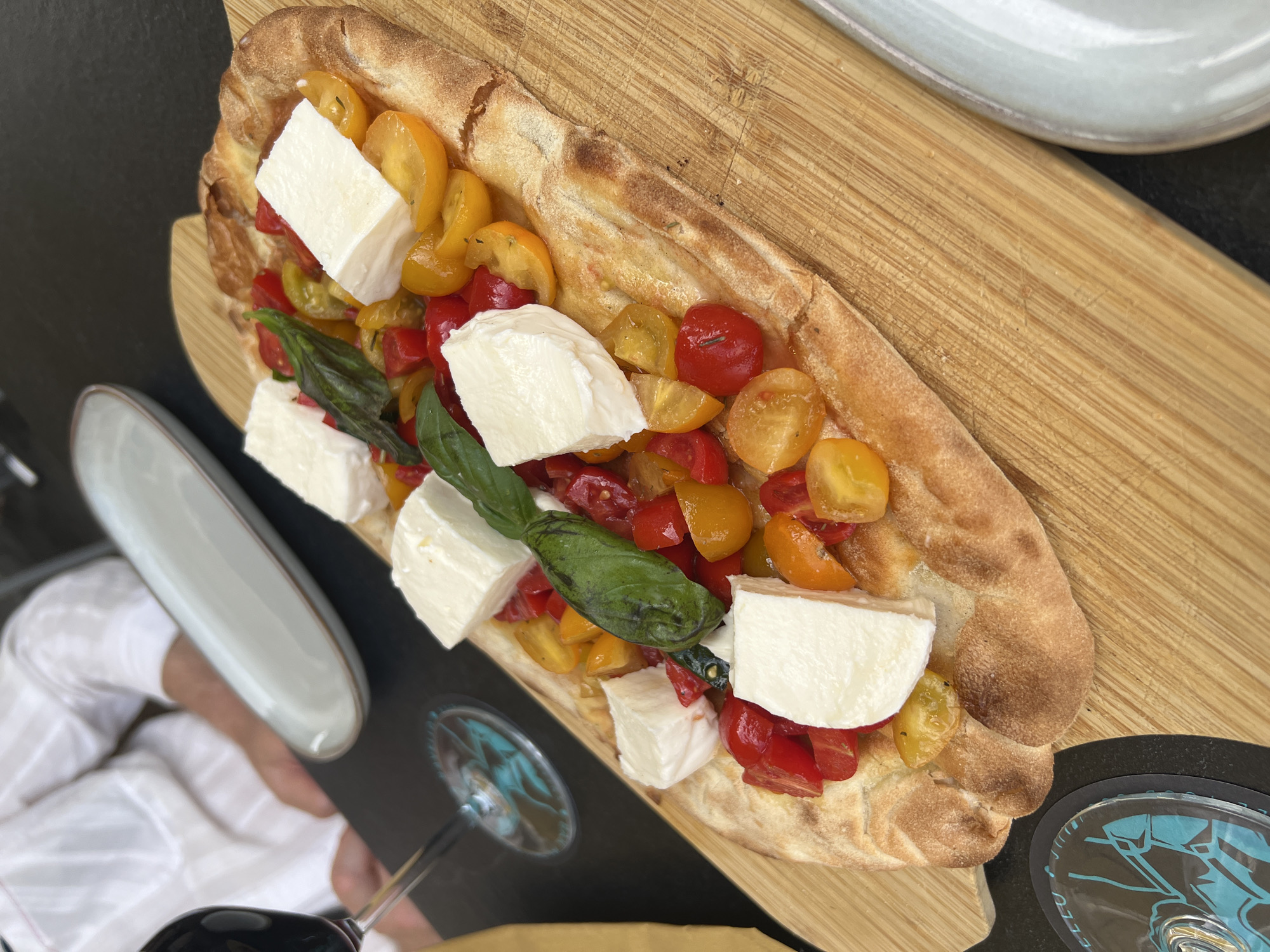 A flatbread pizza topped with fresh mozzarella, cherry tomatoes, and basil leaves is presented on a wooden board. The pizza is oval-shaped with a slightly puffed, golden-brown crust. The mozzarella is cut into rough cubes and scattered across the pizza. Halved cherry tomatoes, in shades of red and yellow, are mixed with the cheese. A few dark green basil leaves are also visible.
The wooden board is placed on a dark surface. To the left, a stemmed glass containing red wine is visible, with its reflection showing a patterned design. In the background, a person wearing a white striped shirt is partially visible. Another white plate with a grey rim is placed behind the pizza.
The scene appears to be in Ravello, Italy, suggesting an outdoor or restaurant setting with a focus on fresh, Mediterranean cuisine. The lighting is natural and diffused, casting soft shadows. The overall impression is one of a delicious and appealing dish presented in a casual yet elegant manner.
