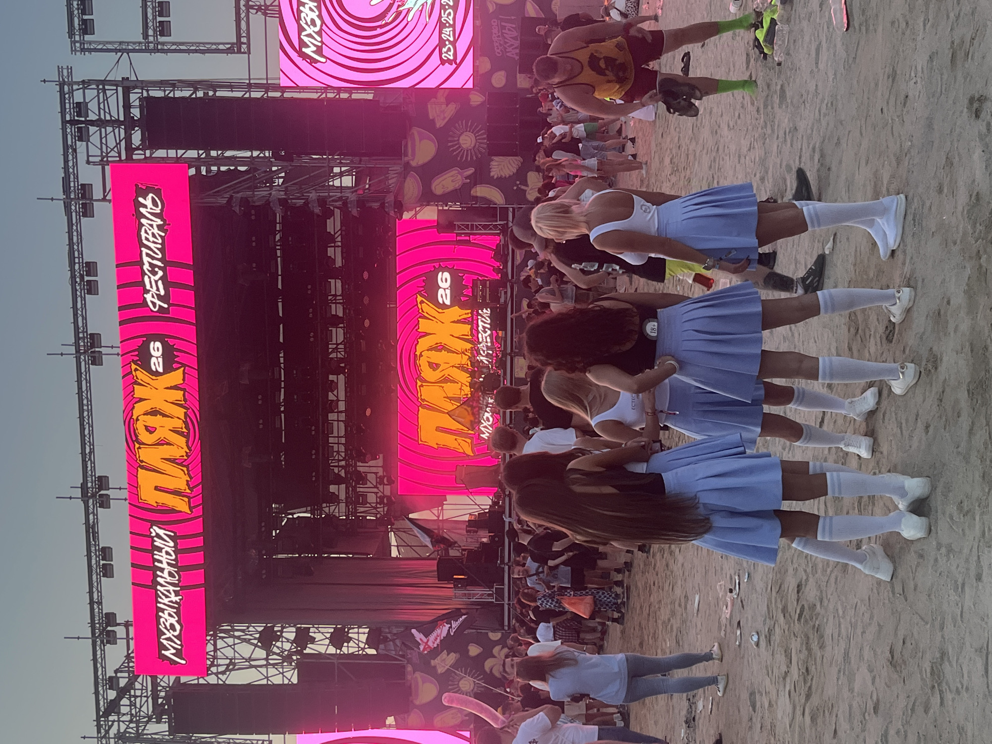This image captures an outdoor music festival, identified as "Яркие Люди" (Bright People) which translates to "Vibrant People" or "Bright Folks" in Surakovo, Russia. The festival appears to be taking place on a beach, with sand covering the ground.
Dominating the scene is a large stage setup with multiple screens displaying the festival's name in bright pink and orange lettering. The top screen reads "МУЗЫКАЛЬНЫЙ ПЛЯЖ 26 ФЕСТИВАЛЬ" (Musical Beach 26 Festival), while a lower screen shows a similar design with "ПЛЯЖ 26" (Beach 26). Various lighting rigs and speaker systems are visible on the stage structure.
A crowd of people, estimated to be in the hundreds, is gathered in front of the stage. They are dressed in casual summer attire, and some appear to be actively watching the performance or socializing. Four young women in the foreground are particularly noticeable, all wearing similar light blue pleated skirts, white knee-high socks, and white sneakers. They are facing away from the camera, towards the stage.
The environment is sandy, with some scattered debris like bottles and paper on the ground, suggesting a well-attended event. The sky is a clear, pale blue, indicating it is likely late afternoon or early evening. The overall atmosphere suggests a lively and energetic festival atmosphere.