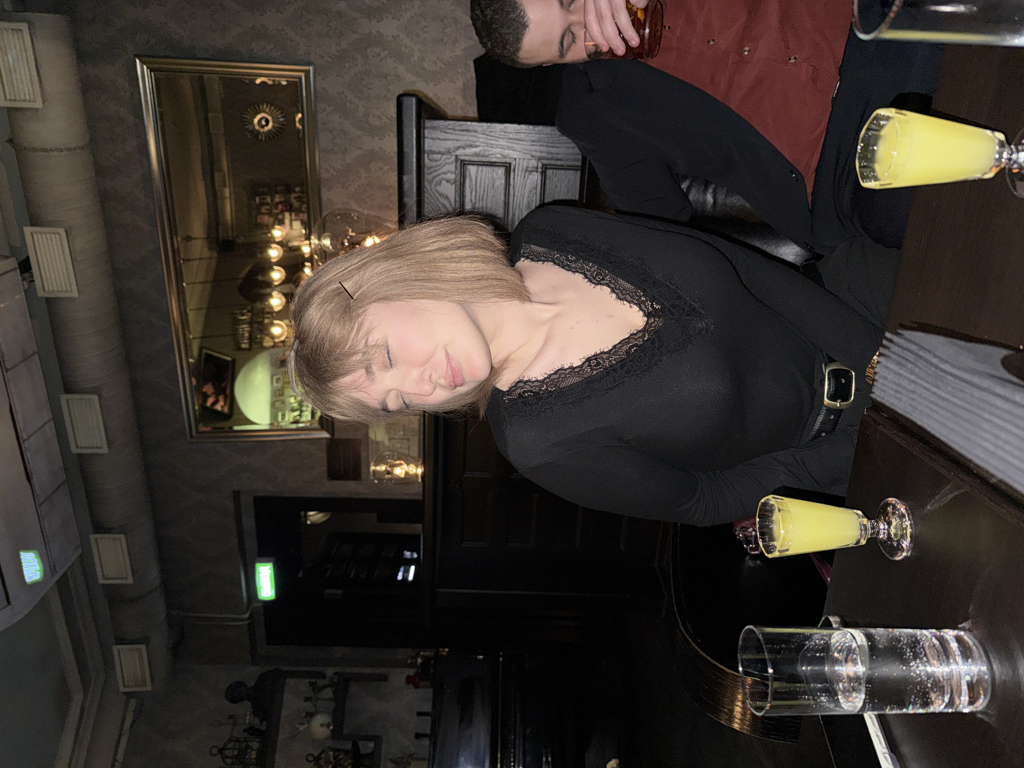 Two people are seated at a dark wooden table in what appears to be a dimly lit bar or restaurant. A woman with light blonde hair and a black top with lace detailing is leaning towards the left, eyes closed, resting her head slightly. Next to her sits a man, out of focus, wearing a maroon or dark red shirt and a black jacket.
On the table are several glasses. One contains a yellow liquid, possibly a shot, and another holds a clear liquid. A partially visible mirror reflects the interior of the establishment, including lighting fixtures and decorative elements.
Based on the architectural features and decor, it is not possible to specifically identify the exact location or confirm that it is in Moscow, Russia.