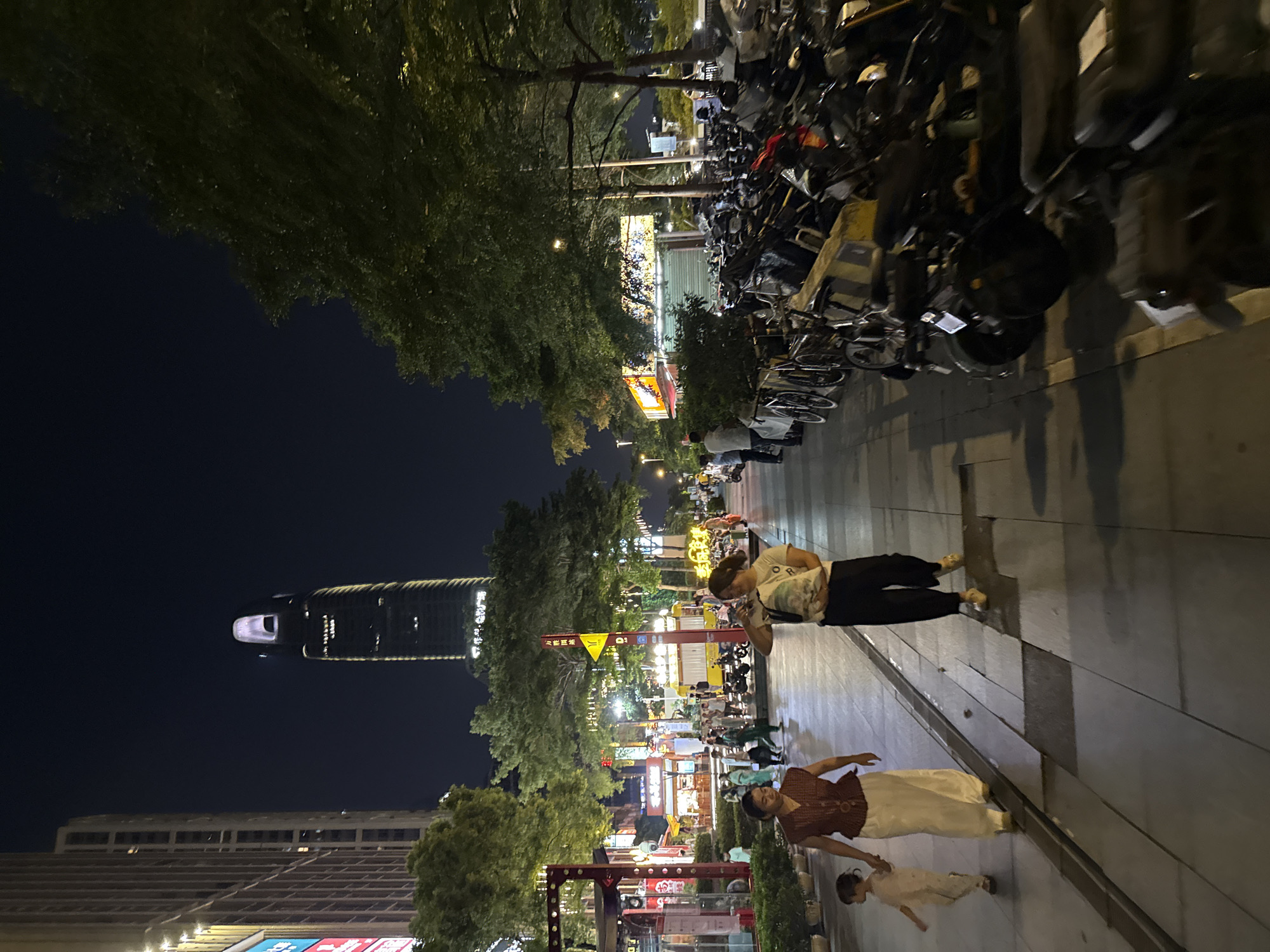 In Haizhu District, China, at night, a sidewalk bustles with activity under artificial lighting. A woman in dark pants and a white top walks towards the camera, holding a patterned bag, and seemingly looking at her phone. Beside her, a woman in a dark red sleeveless top and a long white skirt walks with a young child in a light-colored outfit.
The street is lined with trees and illuminated signs, suggesting a commercial area. A tall, modern skyscraper dominates the background skyline. A row of parked motorcycles and bicycles lines the side of the street. In the distance, food stalls and shops are visible, some with bright lights. The overall scene depicts a lively urban night environment.