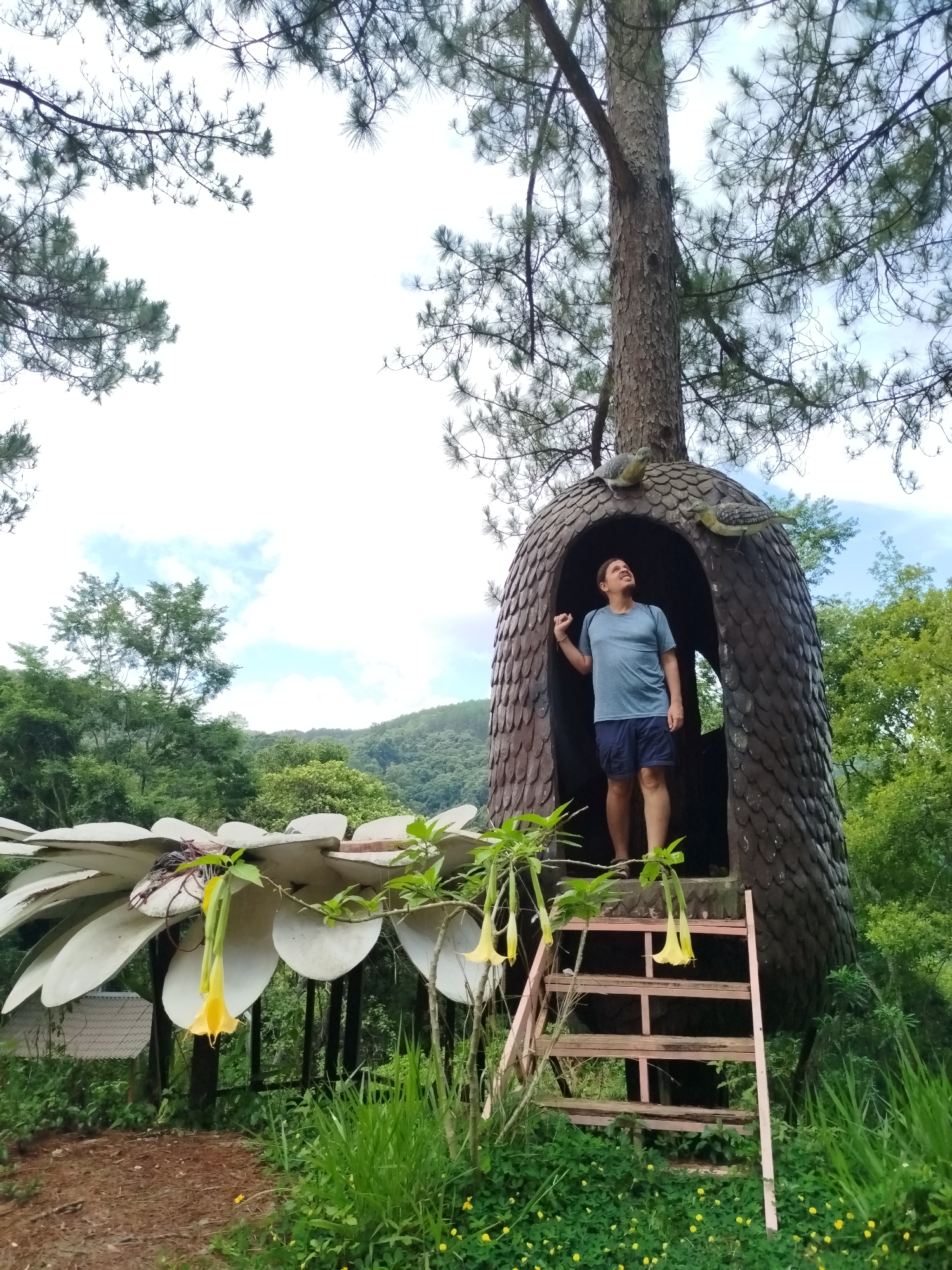 The scene is set in Thôn 5, Vietnam, featuring a fair-skinned man standing inside a treehouse structure. The man is wearing a light blue t-shirt and dark blue shorts. He appears to be looking upwards, with his right hand raised. The treehouse is dark brown and ovoid in shape, built around a tree trunk. Two bird sculptures are attached to the exterior of the treehouse. A wooden ladder provides access to the entrance. To the left, a large artificial flower structure with white petals is visible. Yellow flowers are hanging from the foliage near the treehouse. The surrounding environment consists of lush green vegetation, with trees, grass, and flowers. In the background, a mountain range is partially obscured by trees. The sky is partly cloudy.