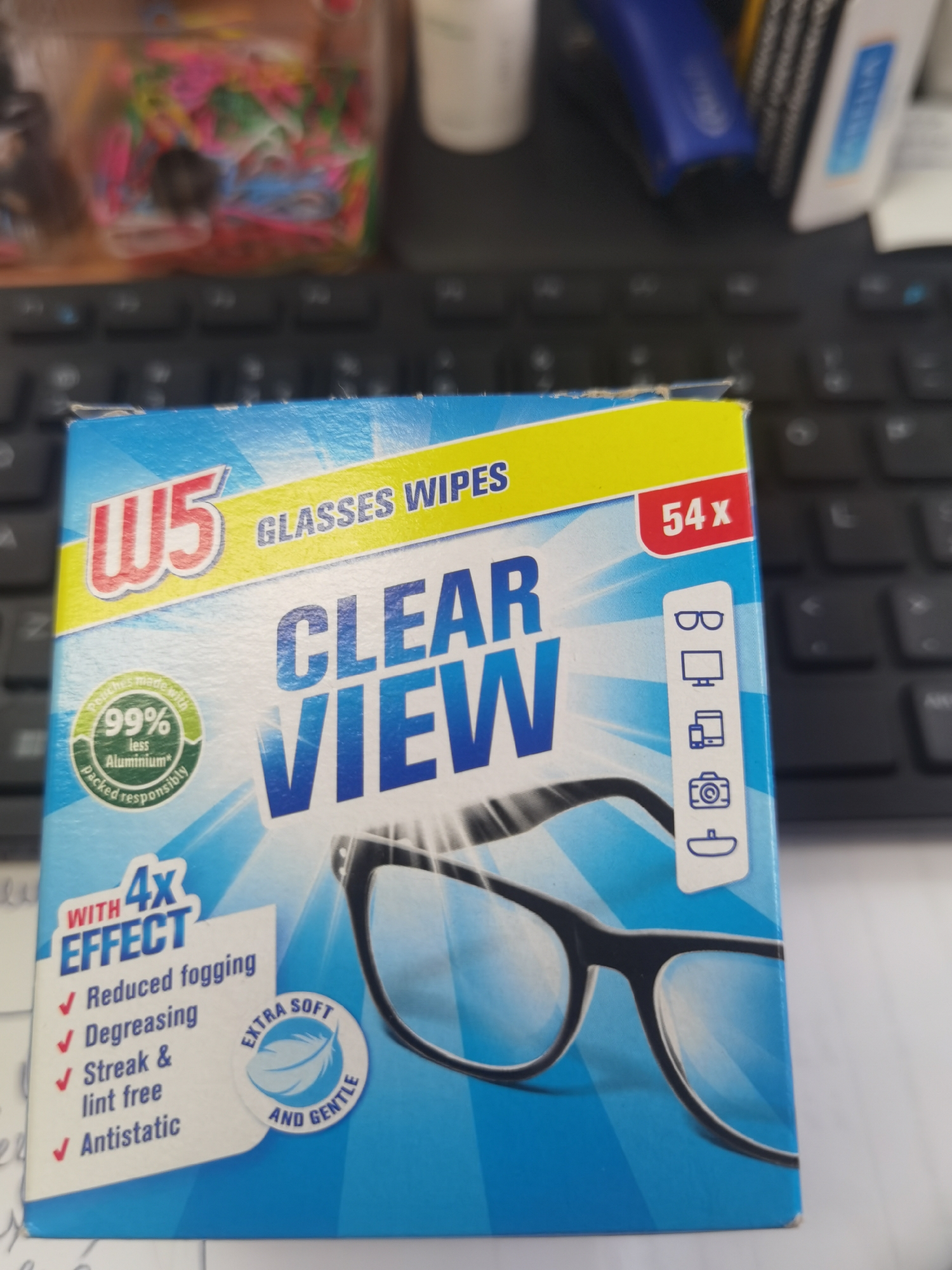 The image shows a close-up of a box of "W5 Clear View Glasses Wipes" placed on a keyboard. The box is predominantly blue with yellow and red accents. The branding "W5" is prominent in red and white, accompanied by the text "GLASSES WIPES" in yellow. The words "CLEAR VIEW" are printed in large blue letters across the center of the box. To the right, a red rectangle indicates "54x". On the left side of the box, there is a circular green seal stating "Pouches made with 99% less Aluminium packed responsibly". Below this, text indicates "WITH 4x EFFECT" and lists benefits: "Reduced fogging", "Degreasing", "Streak & lint free", and "Antistatic". An emblem on the right side features icons for glasses, a computer monitor, a mobile phone, and a camera, suggesting the product's versatility. A circular emblem with a feather graphic states "EXTRA SOFT AND GENTLE". The background consists of a black computer keyboard, suggesting an indoor setting. There is no indication of the city of Lomets, Bulgaria, in the image.