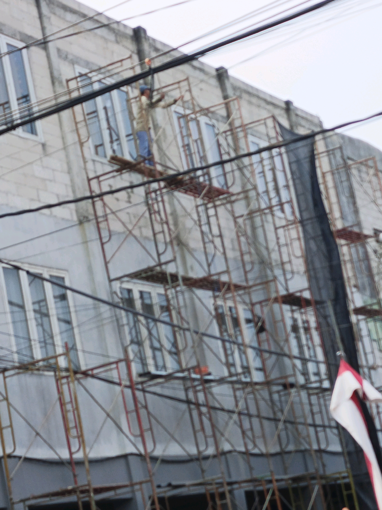 In Ketegan, Indonesia, a construction worker, wearing a light-colored long-sleeved shirt and blue pants, is positioned on scaffolding. He is facing right, with his arms extended outwards as if engaged in work or signaling. The scaffolding, constructed from metal poles with red and orange accents, surrounds a multi-story building. The building's exterior features light-colored brickwork on the upper levels and is punctuated by numerous windows with white frames. A black mesh screen is visible on the right side of the scaffolding. Several electrical wires are strung across the foreground, obscuring parts of the scene. A flag, predominantly white with red and black stripes, is partially visible in the lower right corner of the image, attached to a pole extending from the scaffolding. The sky is overcast, suggesting a cloudy day.