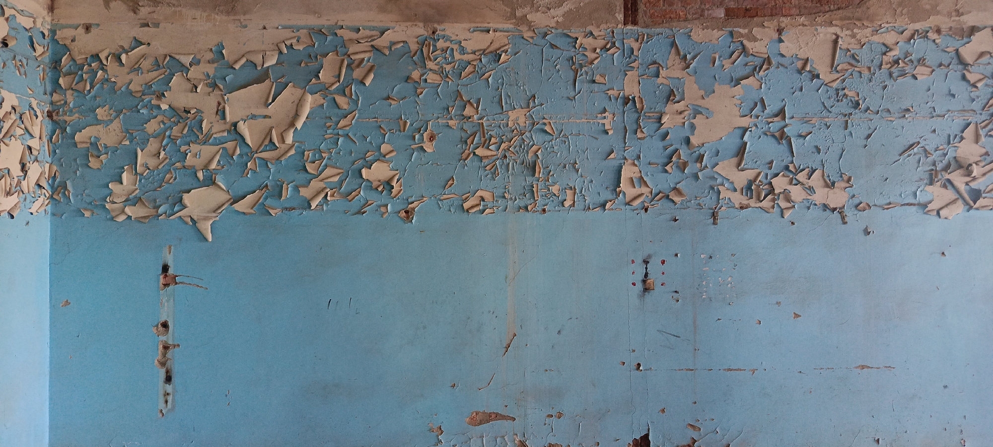 The image shows a close-up of a dilapidated interior wall, likely in an abandoned building. The wall is painted a faded, pale blue and is heavily textured with peeling, chipped paint in shades of cream and beige. The upper portion of the wall exhibits significant damage, with large sections of the top layer of paint flaking away to reveal the underlying blue and some reddish-brown material, possibly brick or plaster, beneath. There are several vertical marks and holes on the blue section of the wall, suggestive of previous fixtures or damage. The overall impression is one of decay and neglect. The location is stated to be in the city of Baku, Azerbaijan.