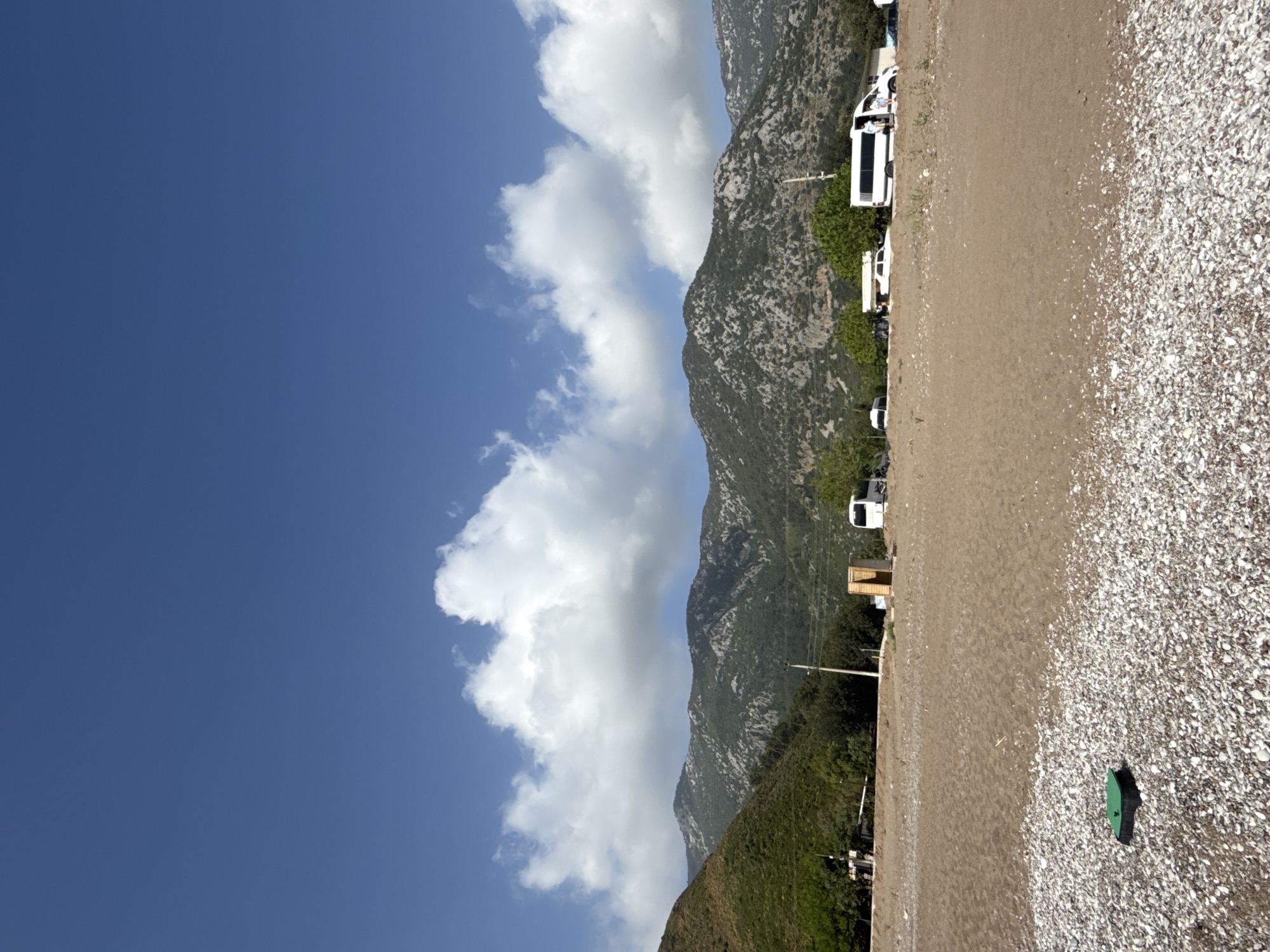 The image depicts a landscape in Kumluca, Turkey. A mountain range with rocky slopes covered in sparse vegetation runs parallel to a beach composed of sand and small white pebbles. White clouds partially obscure the mountains against a bright blue sky. The coastline is lined with a row of white vehicles, possibly vans or small buses. At the edge of the beach is a green object of ambiguous purpose.