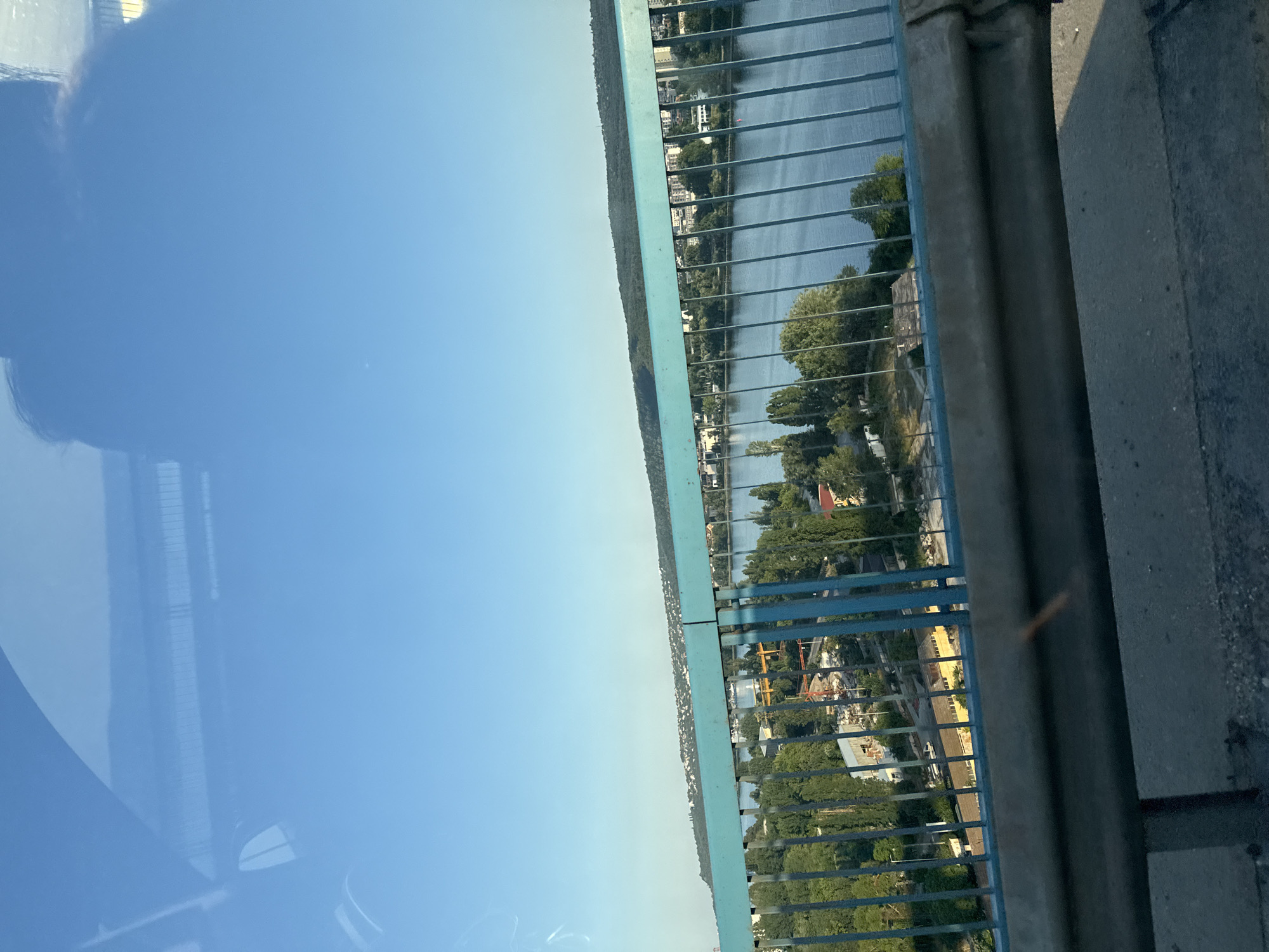 An eye-level, medium outdoor shot captures a view from a bridge on a clear, sunny day. The sky is a bright blue, with a subtle reflection of a person's head and shoulders visible in the upper left quadrant, likely from the windshield of a vehicle. A turquoise blue metal railing runs horizontally across the foreground, with vertical bars spaced evenly.
Below the railing, a body of water, possibly a river or lake, stretches into the distance. The water is calm and reflects the blue sky. On the far bank, a densely populated city is visible, with buildings of various sizes and colors nestled amongst green trees. A distant, forested hill forms the horizon line.
In the middle ground, on the bank of the water, there are more trees and some buildings, including what appears to be a yellow, rectangular structure. Hints of construction activity, with a red crane, are also present. The water surface has a slight ripple. The overall scene suggests an urban environment with a significant natural element.