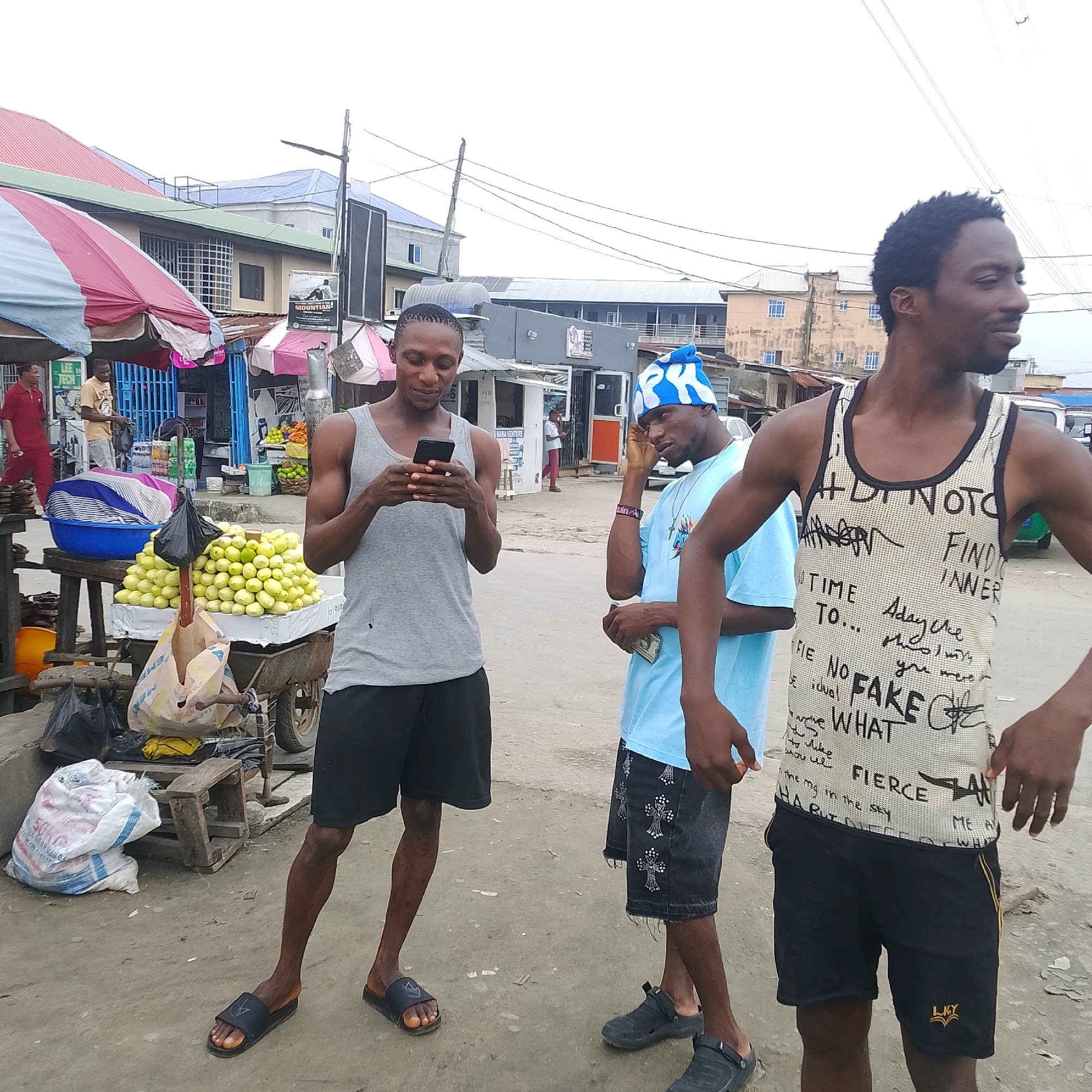 Three young men are standing on a dirt road in what appears to be an outdoor market in Ado, Nigeria. The man on the left, wearing a grey tank top and black shorts, is looking down at his phone. The man in the middle, wearing a light blue t-shirt and dark shorts with a blue and white patterned beanie, is touching his head. The man on the right, wearing a white tank top with black text and black shorts, is looking to his right. In the background, there are market stalls with umbrellas, produce, and various goods. Buildings, power lines, and a cloudy sky are also visible. The ground is dusty and uneven.