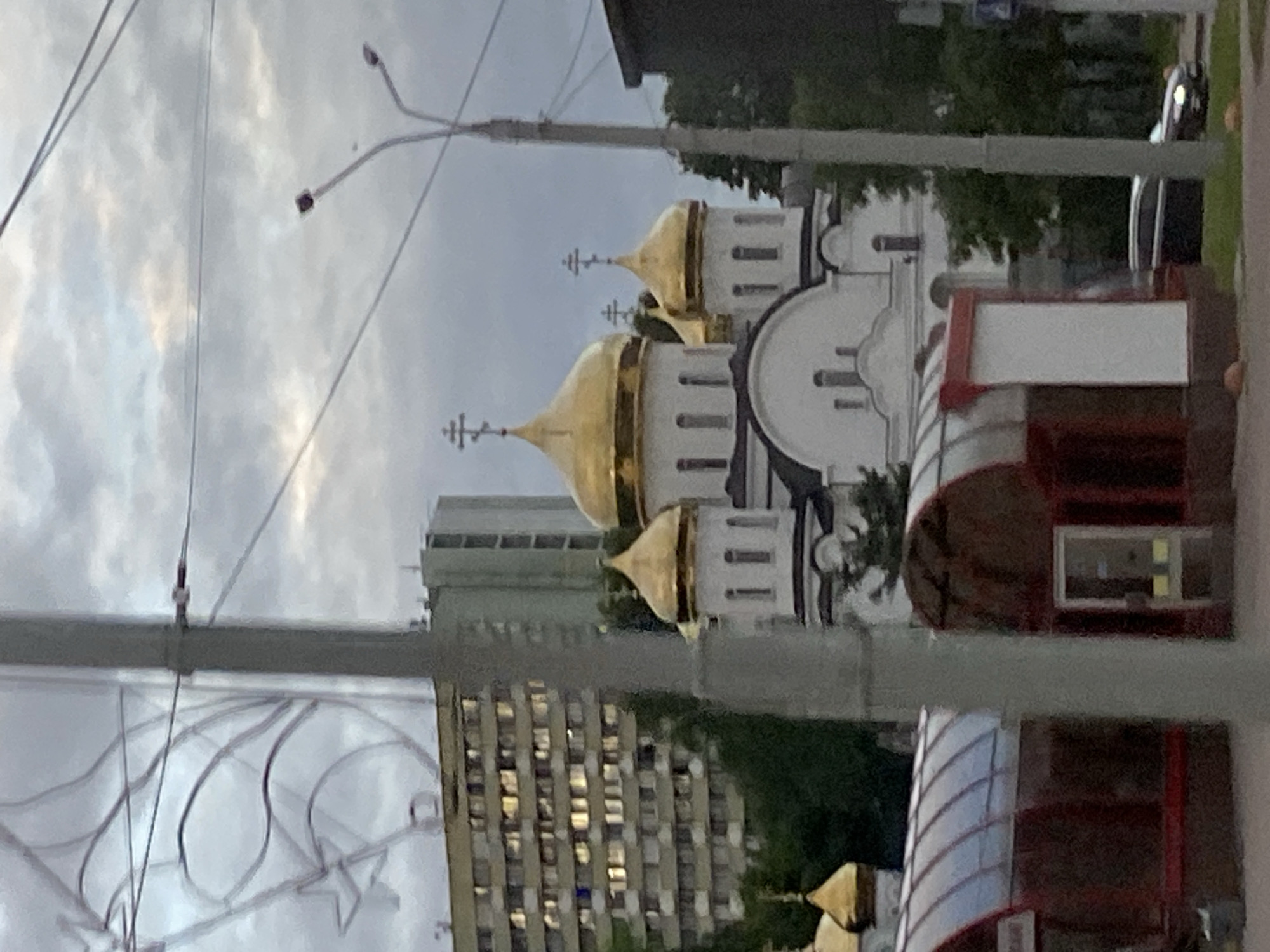 The image depicts an outdoor scene dominated by a white building with multiple golden domes, characteristic of an Orthodox church. The building's architecture features arched windows and decorative elements. In the foreground, a street light pole with wires stretches across the frame, obscuring parts of the view. A modern building with numerous windows, reflecting the light, stands behind the church. Trees and other greenery are visible in the background, suggesting an urban or suburban setting. The sky is overcast, indicating a cloudy or possibly rainy day. The overall impression is that of a community gathering place or a religious site within a developed area. The image appears to be taken during daylight hours, likely late afternoon or evening given the lighting conditions. No people are clearly visible, but the presence of a building suggests it is a public space. There is no visible text in the image.